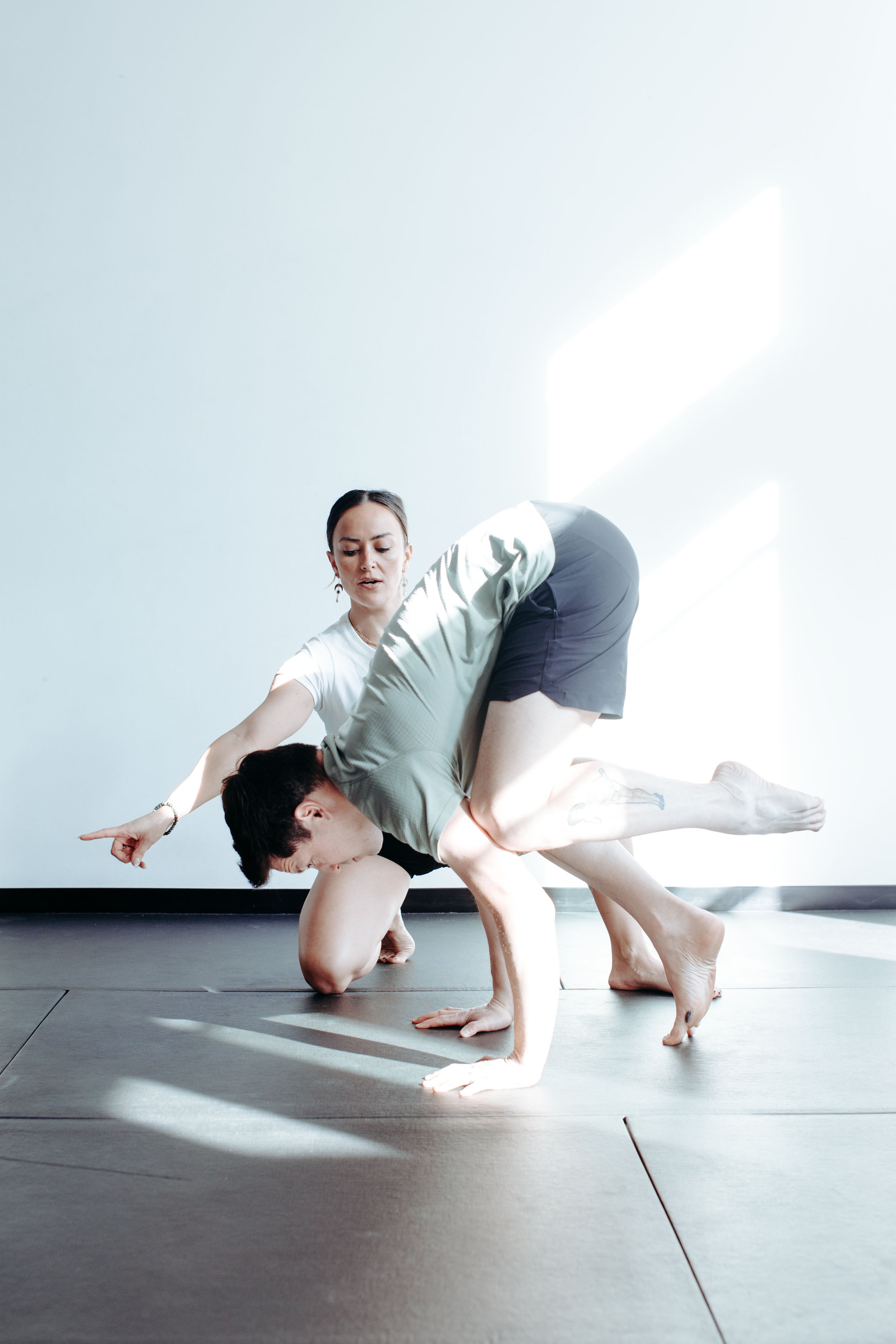 Two people practicing acro yoga inside a room with white walls and sunlight streaming in.