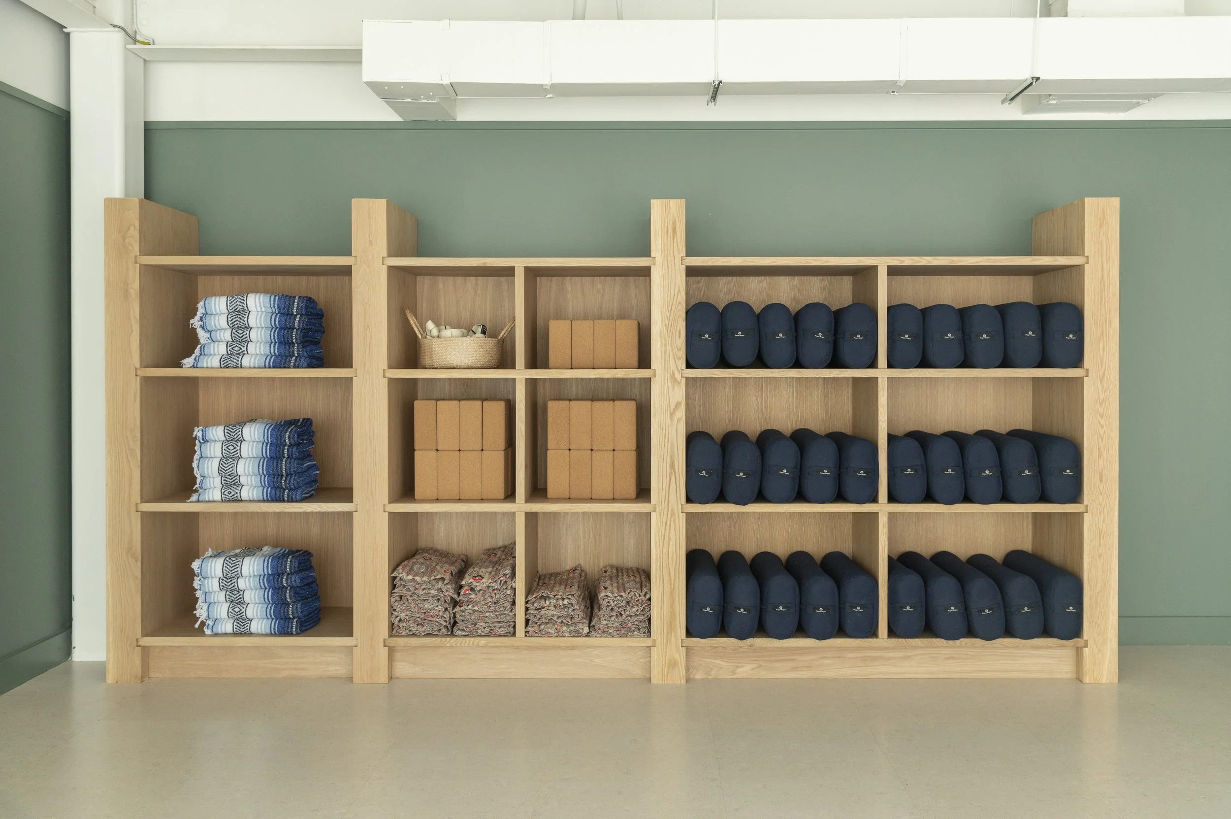 Interior of a fitness or yoga studio with wooden shelving and display of water bottles and athletic wear, modern artwork on the wall, and a tall vase with dried grass by the window.
