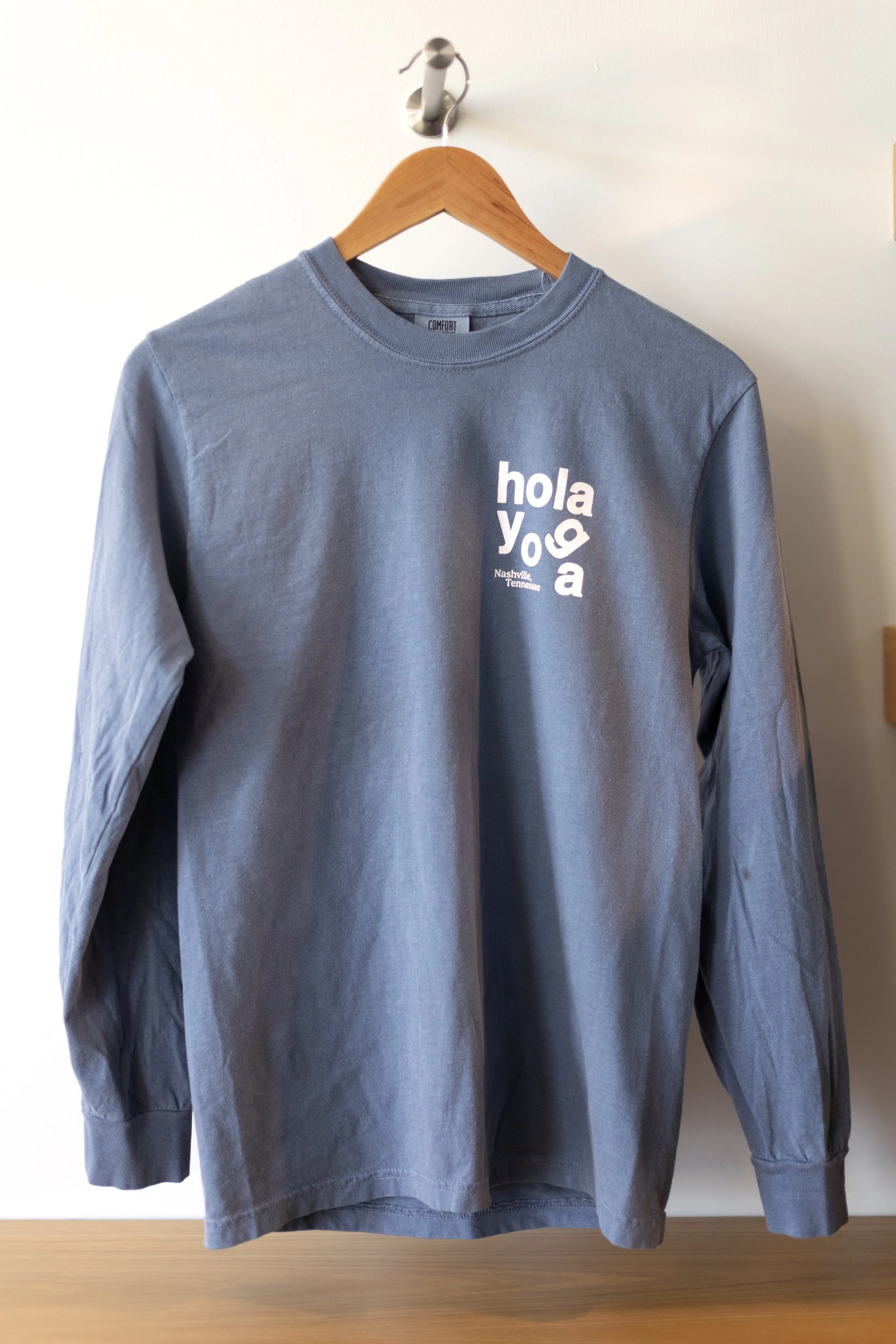 Gray long-sleeve shirt hanging on wooden hanger on a wall with text 'hola yoga Nashville Tennessee' printed on the chest.