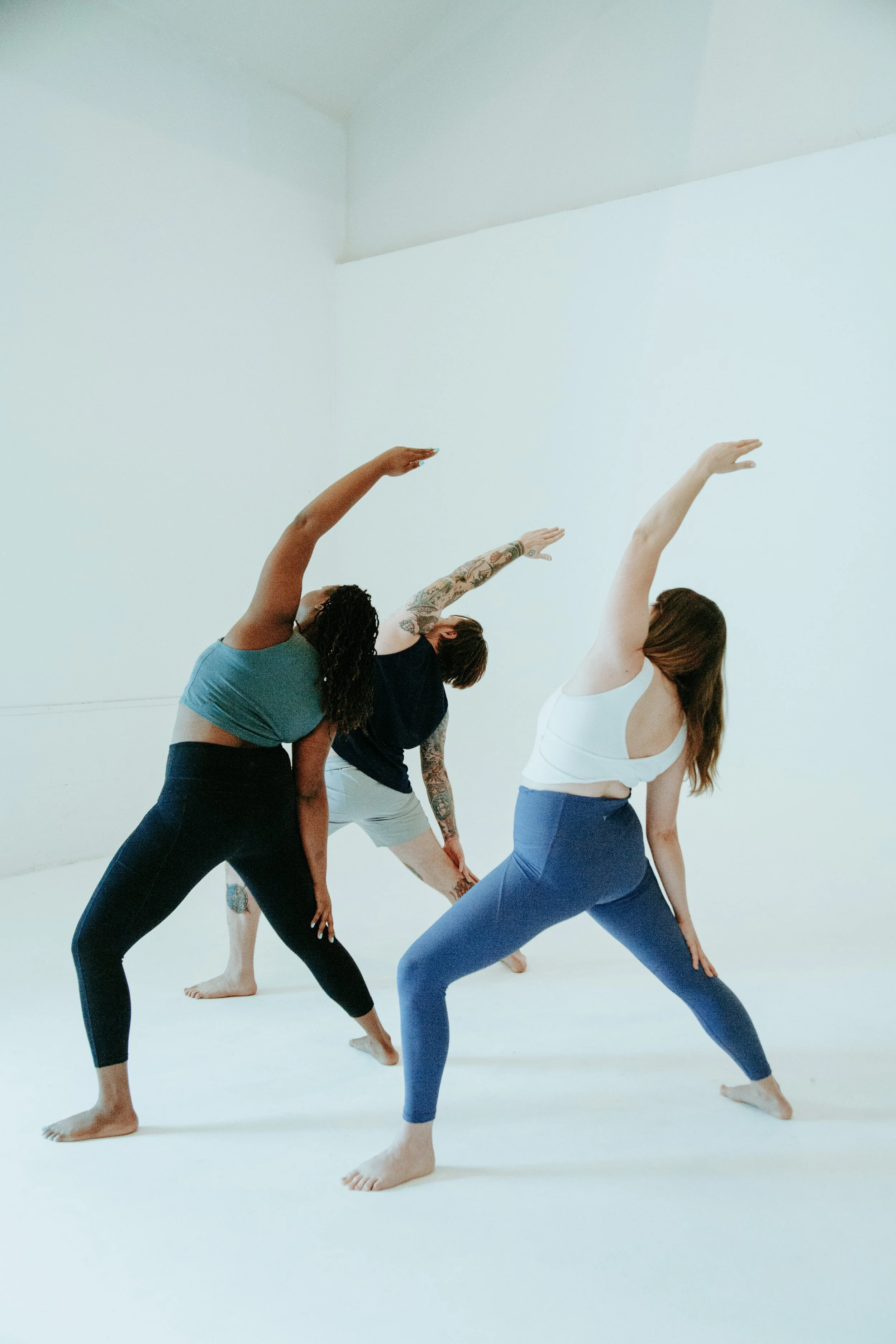 Three women practicing yoga indoors, stretching in a side bend pose with one arm extended overhead and the other reaching down toward their legs.
