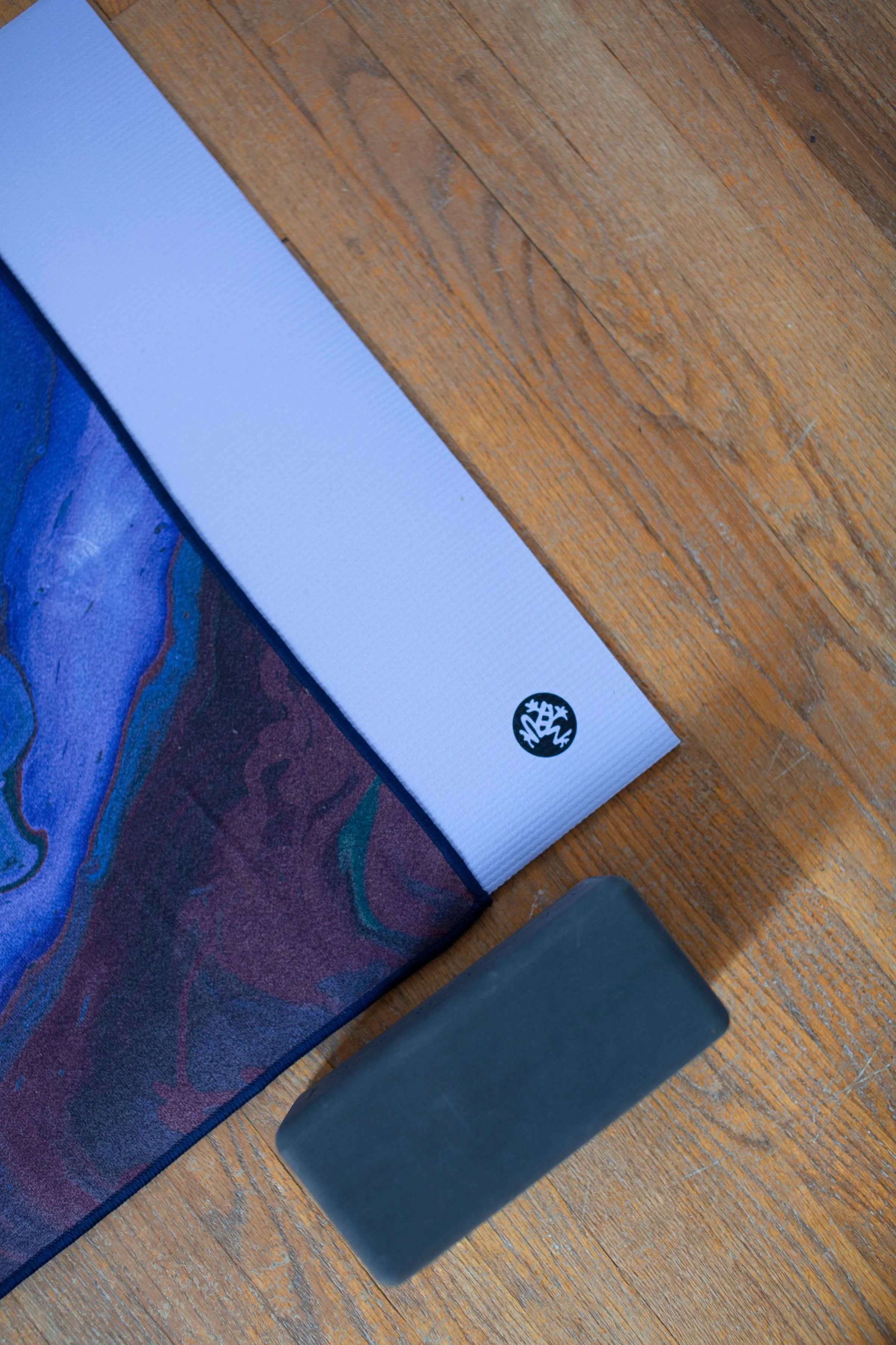 A section of a yoga mat with a vibrant, colorful design, a white foam roller with a black logo, and a black rectangular object on a wooden floor.