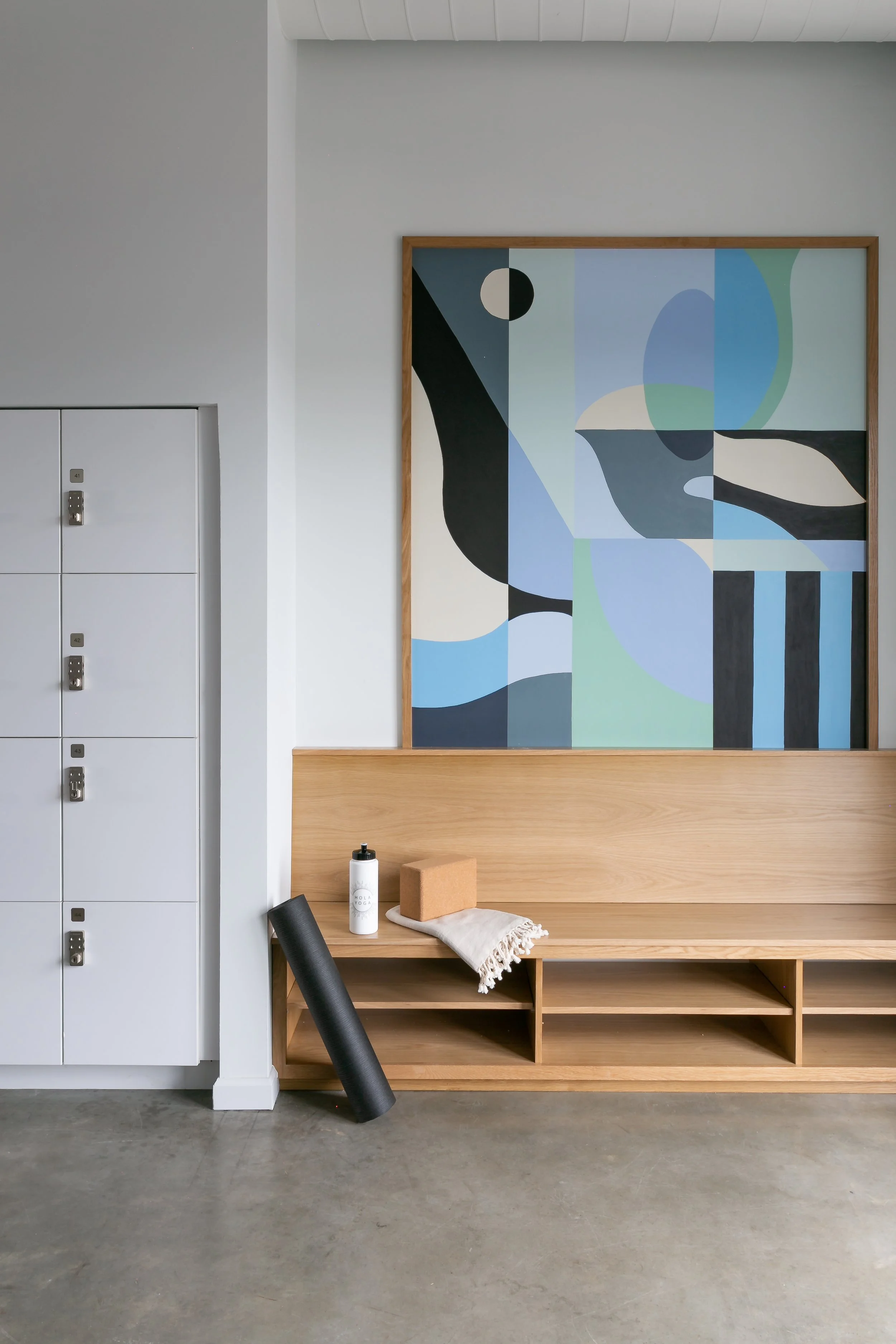 Interior of a minimalist room with a wooden bench, a white water bottle, a cork yoga block, a rolled-up black yoga mat, and a beige cloth on the bench. A large abstract geometric painting in shades of blue, black, white, and green hangs on the wall behind the bench. To the left, there are white lockers with silver locks.