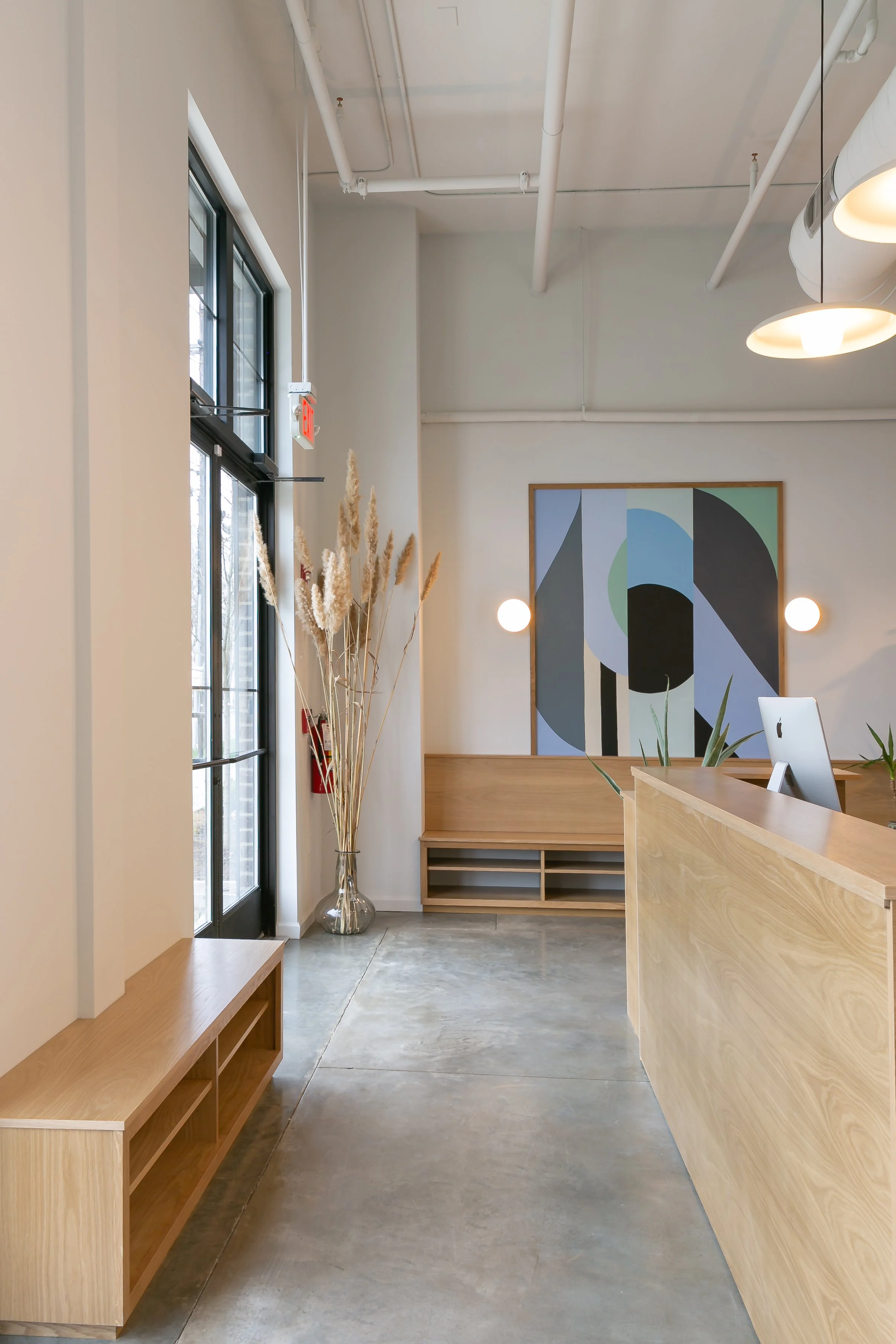 Modern interior space with a wooden reception desk, a large abstract artwork on the wall, and a tall glass vase with dried pampas grass near a window.