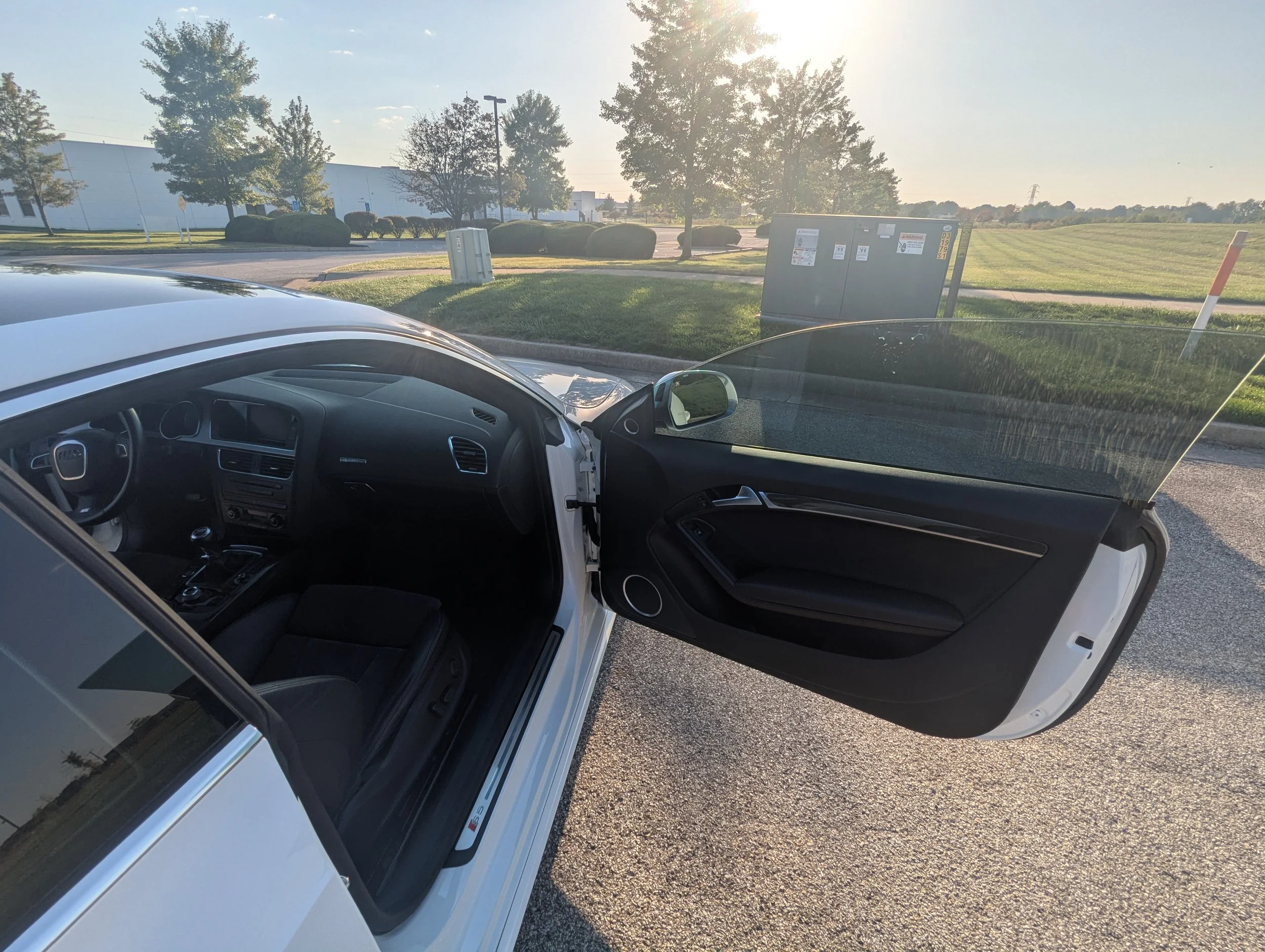 A white car with an open driver's side door parked in a lot during sunset, with green grass, trees, a utility box, and a background of fields and clear sky.
