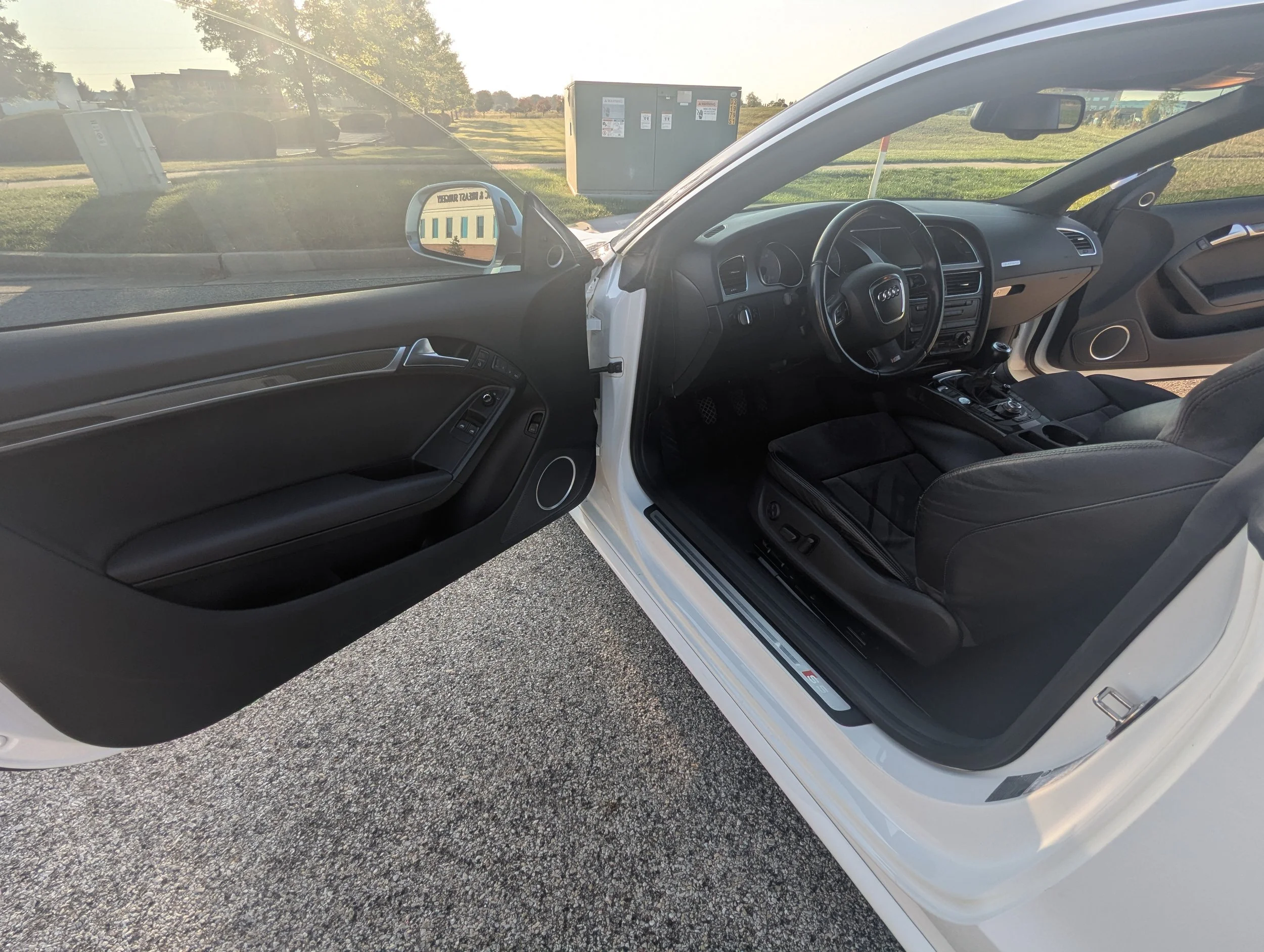Open car door revealing black interior and dashboard of a white car parked outdoors with grass and utility boxes in background.
