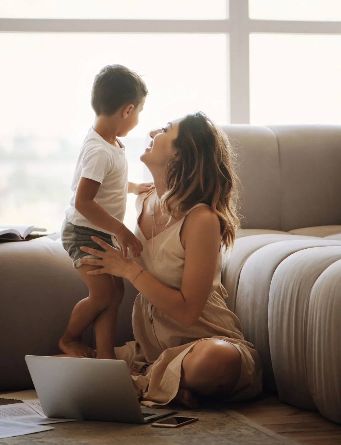 Woman smiling at young child by a sofa, sunlight streaming in, laptop and papers from virtual therapy session on the floor nearby.