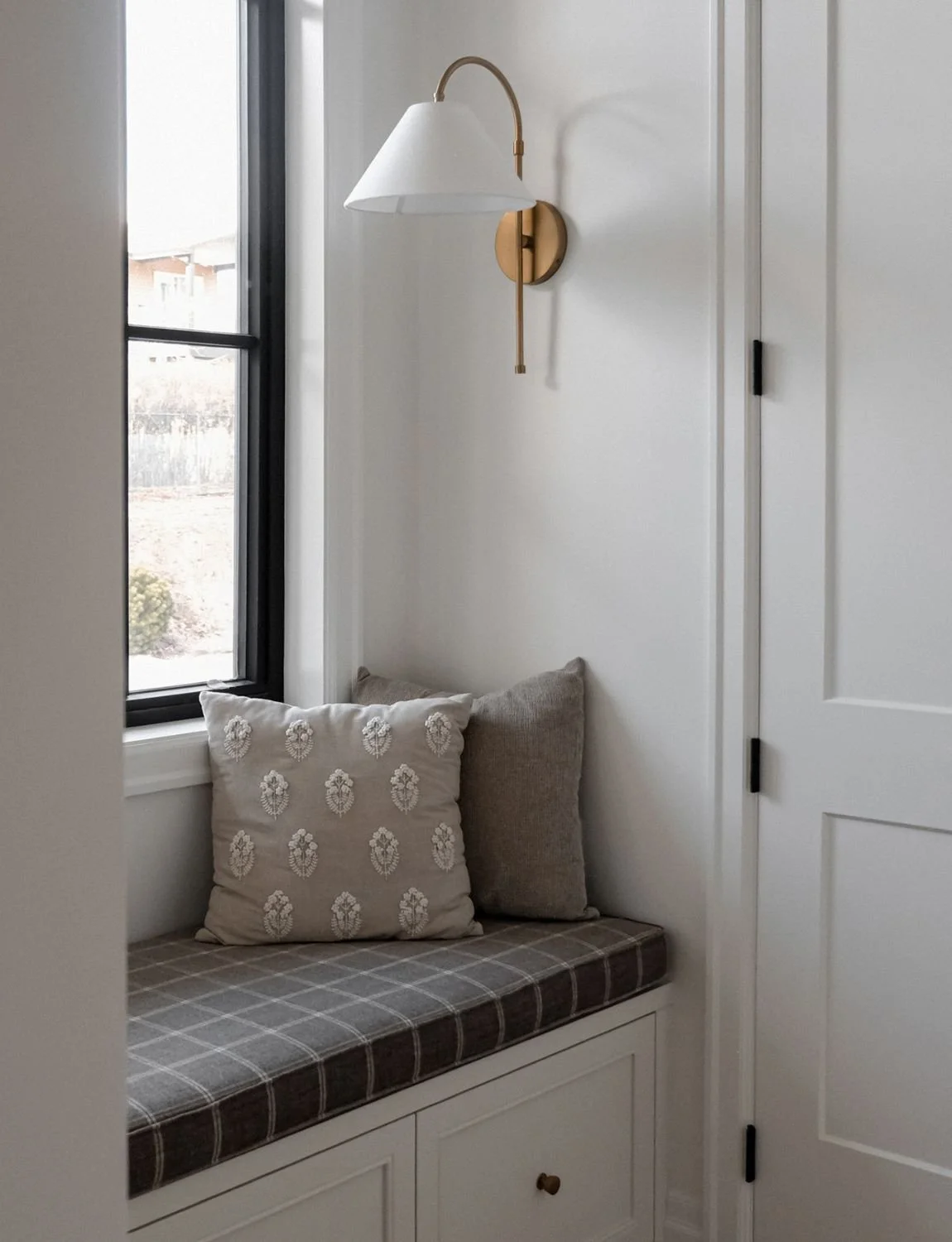 A cozy window seat with two cushions, ideal for a Minneapolis therapist office, under a lamp by a black window and white door.