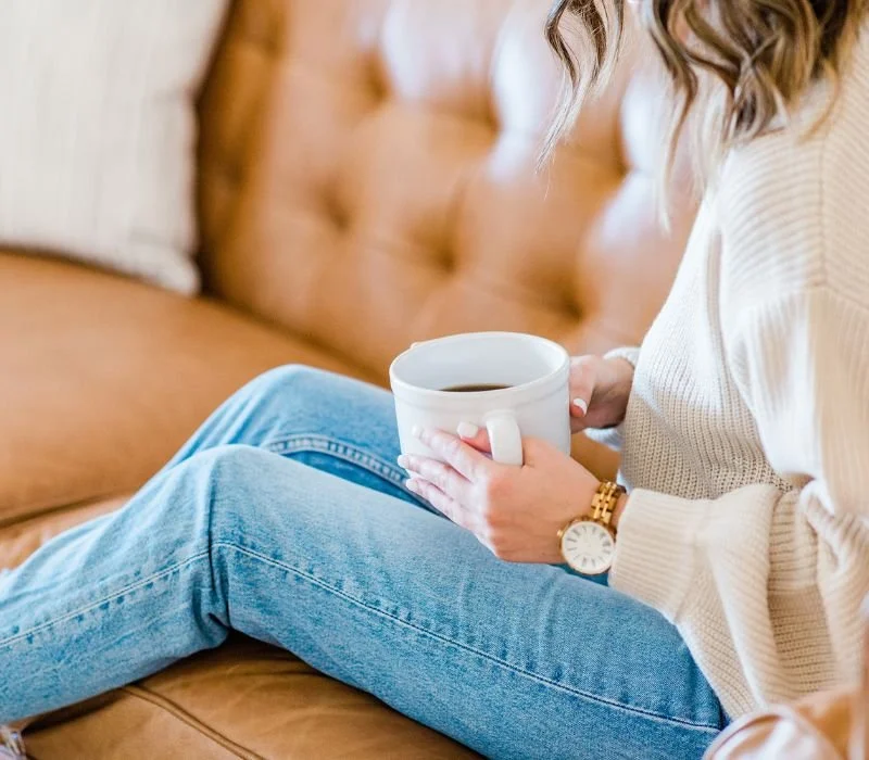 Person sitting on a tan couch, holding a coffee mug, wearing jeans and a cream sweater, enjoying a moment of therapy and relaxation.