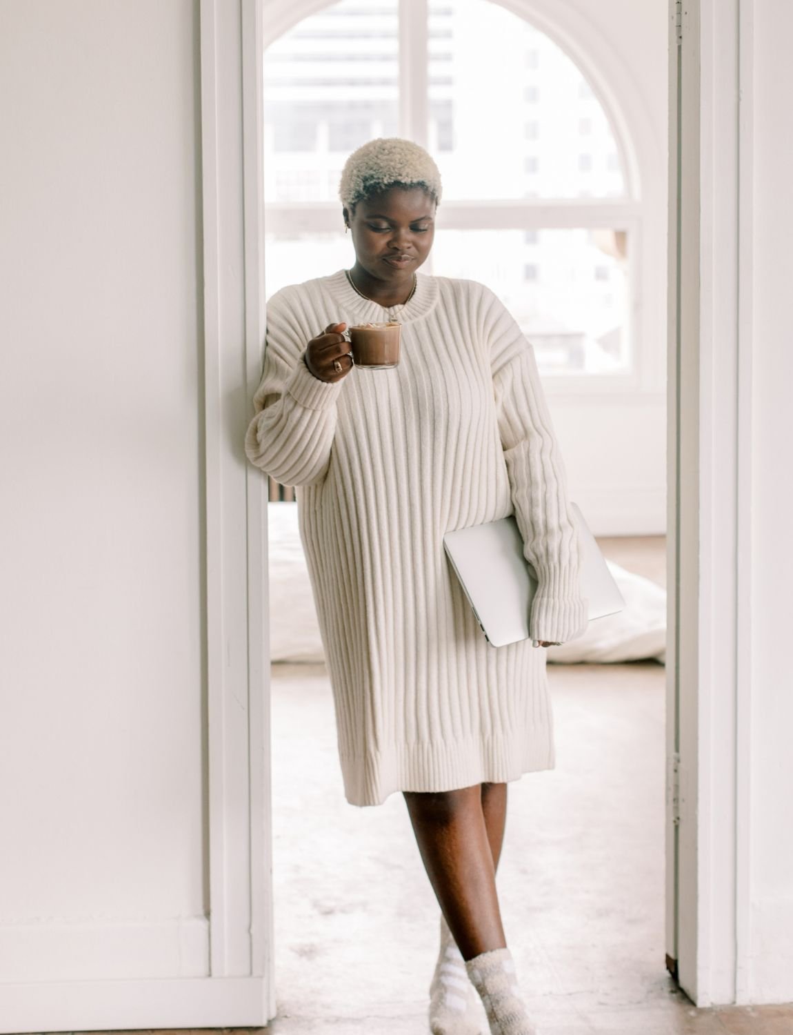 A black therapist in Minneapolis, MN a cream sweater dress stands in a doorway, holding a mug and closed laptop, soft light streaming from a large window.