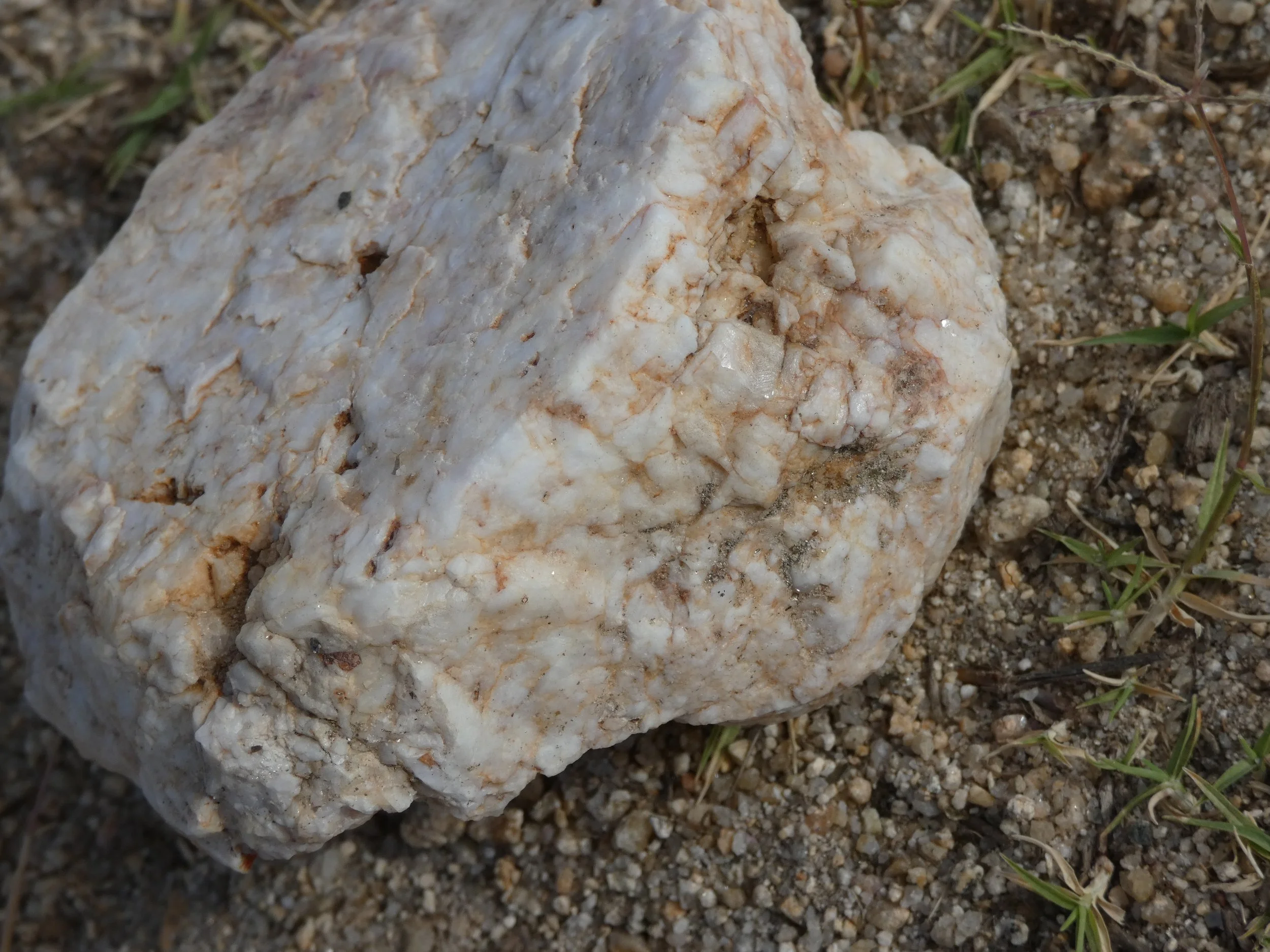 Close-up of a large, white, irregularly-shaped rock with orange and brown streaks, lying on sandy ground with small pebbles and some sparse grass.