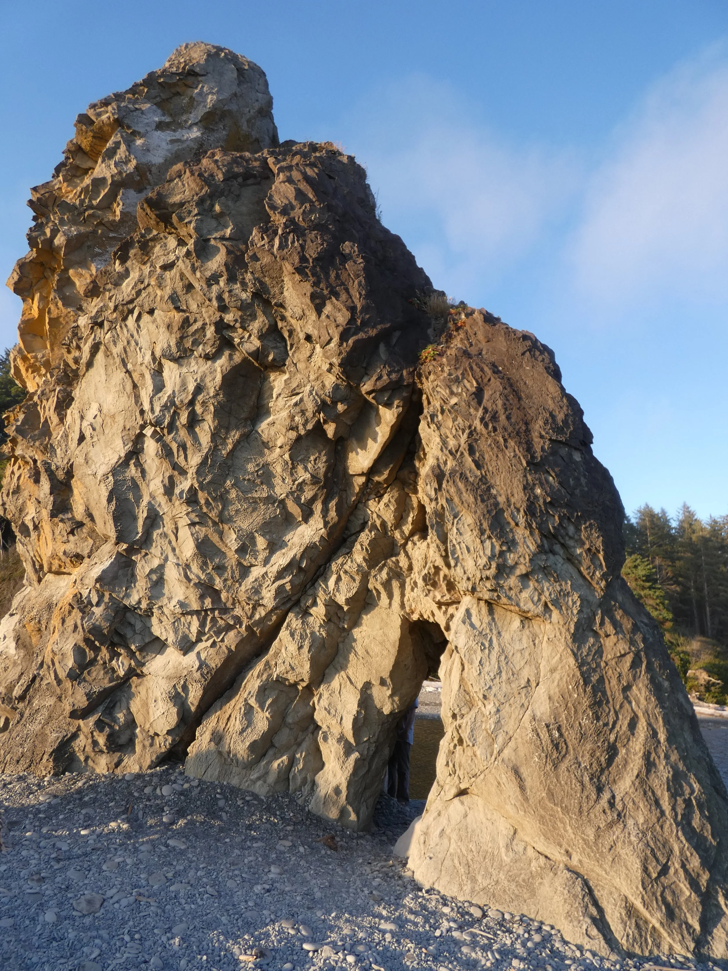 Large natural rock formation with multiple boulders stacked together outdoors, bright blue sky, and sparse vegetation in the background.