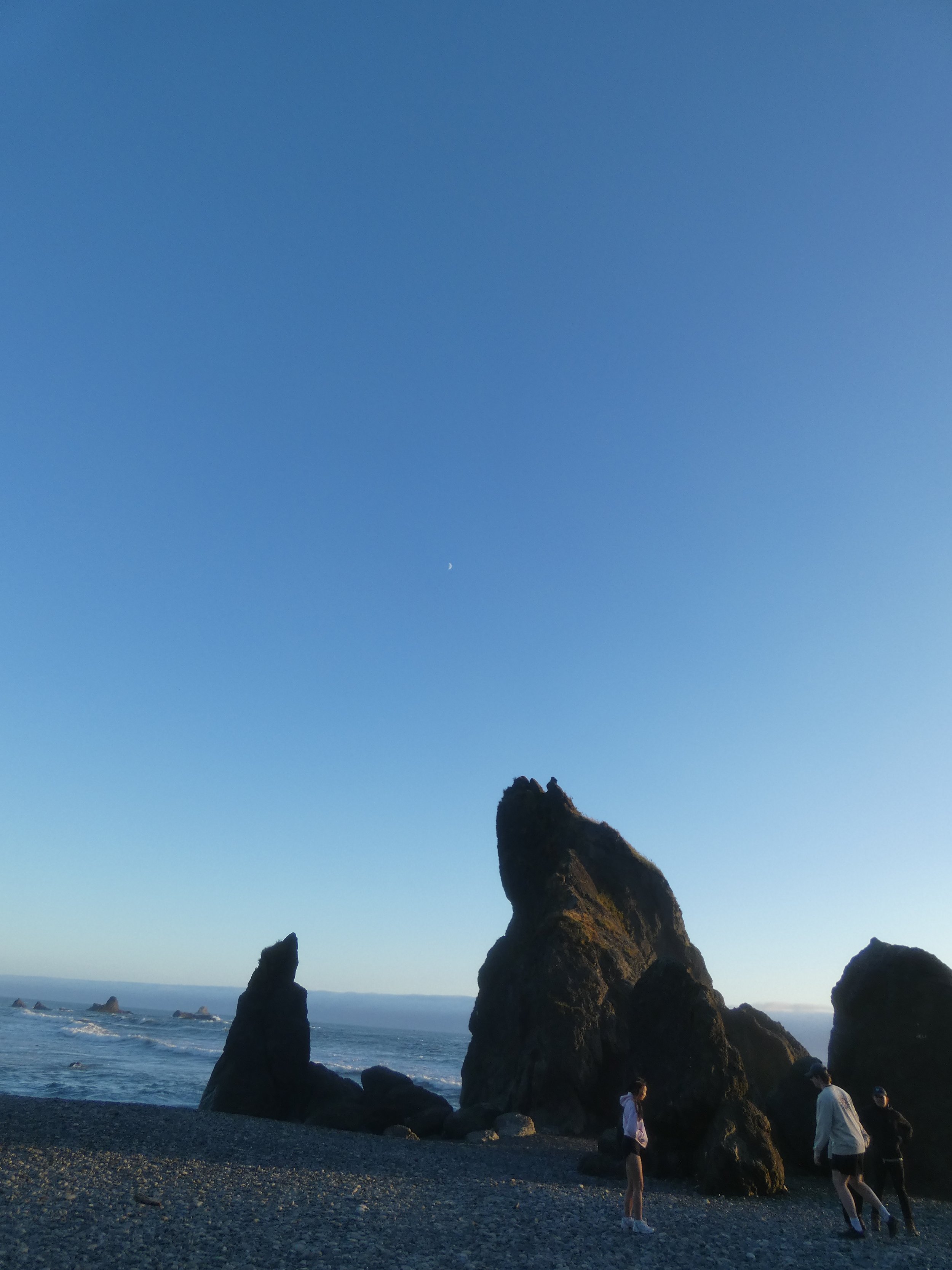 A beach with large rock formations in the ocean and a clear sky with a small crescent moon visible.