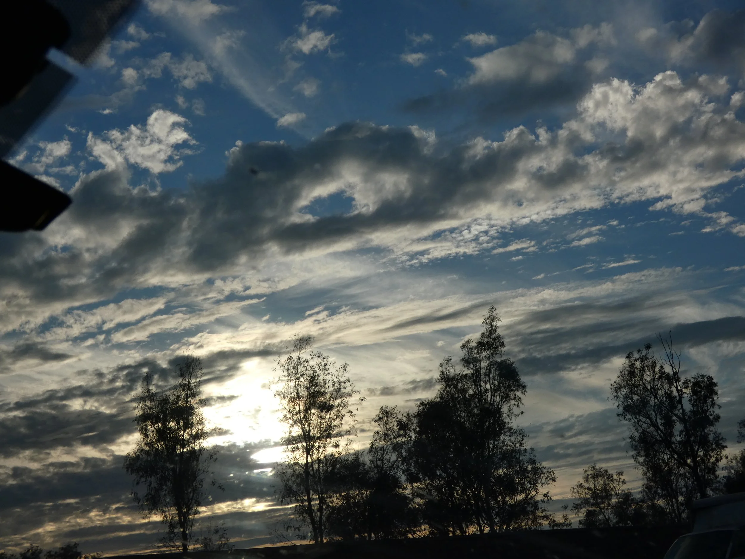 The photo depicts a cloudy sky during sunset or sunrise with the sun partially obscured by clouds. Silhouettes of trees are visible in the foreground.