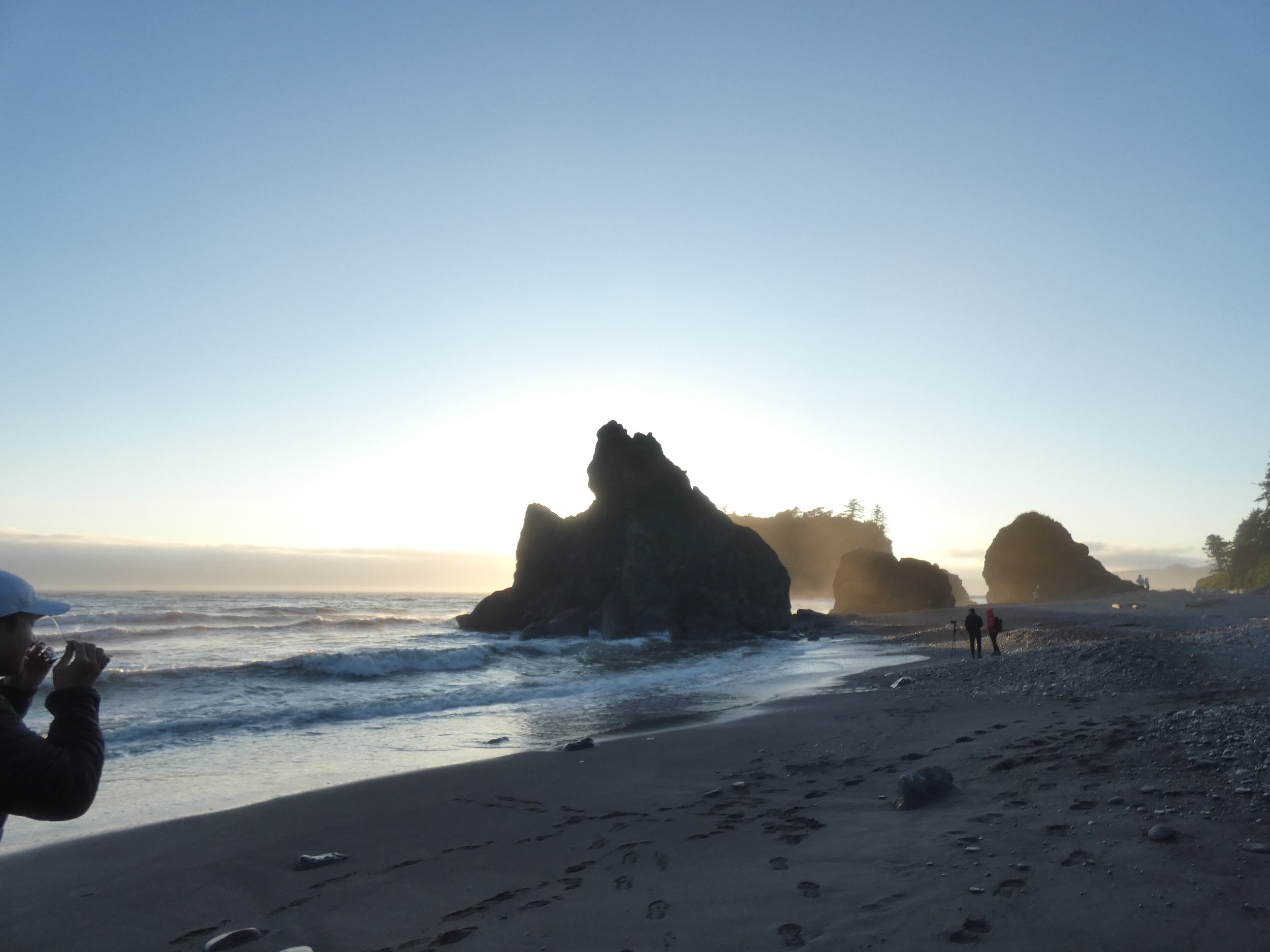 Sunset over a rocky beach with large sea stacks, people taking photos, and a silhouette of trees on distant cliffs.