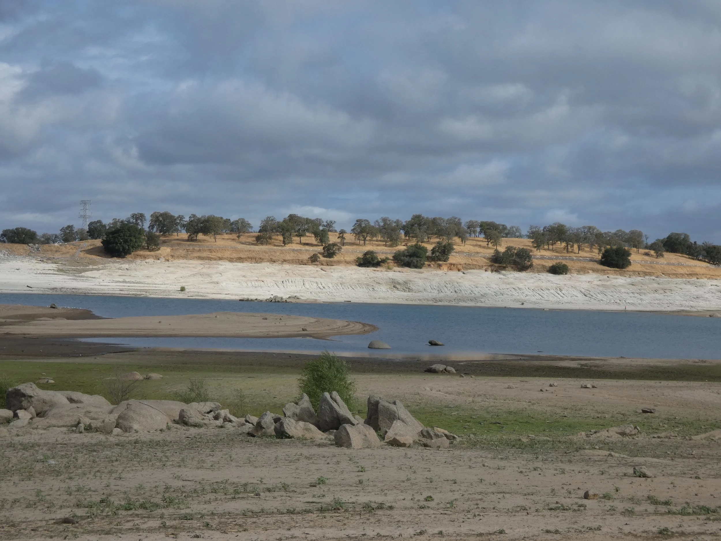A landscape with a body of water in the foreground, dry grass and scattered rocks, a hill with numerous trees in the background, and a cloudy sky overhead.