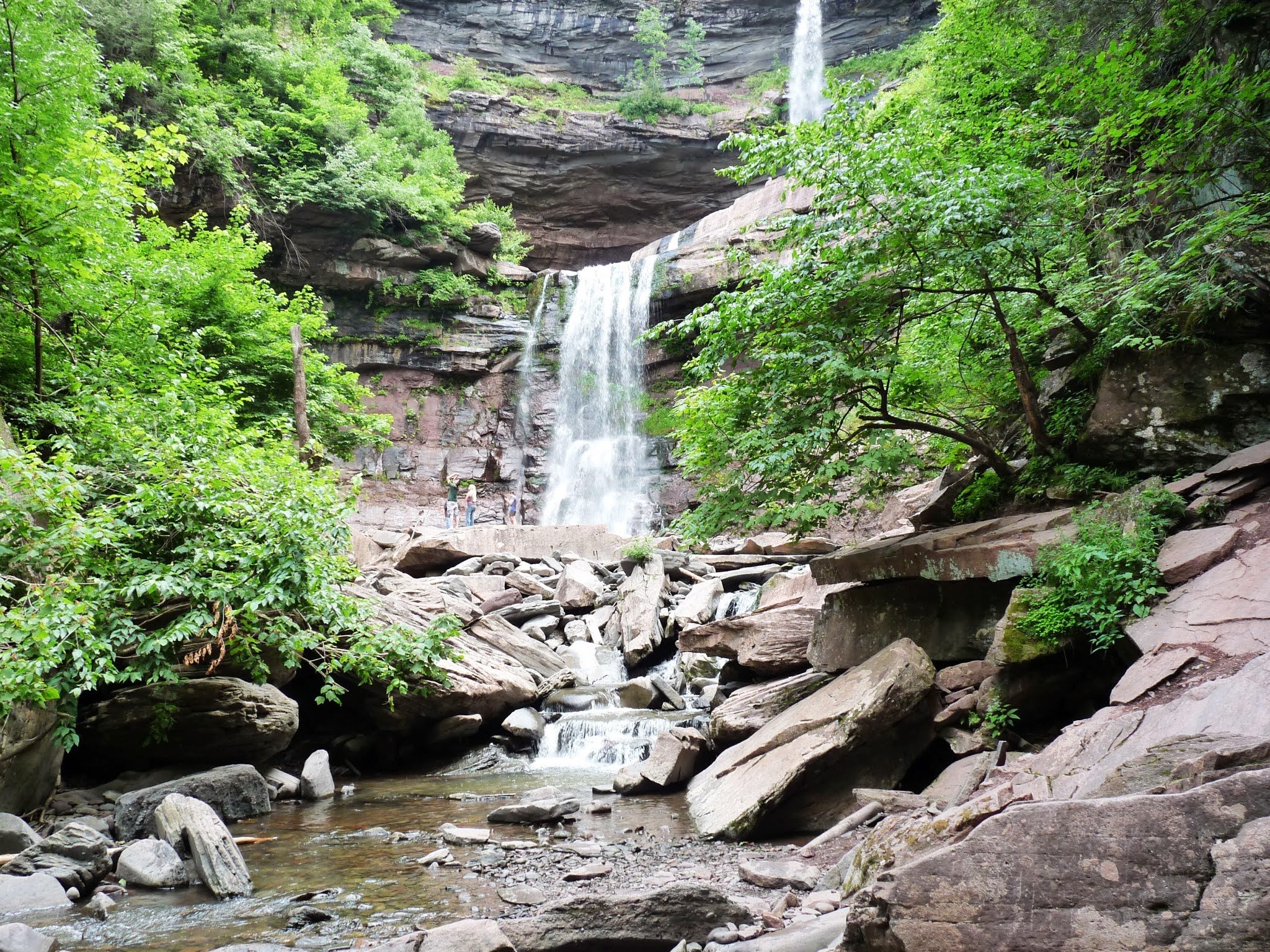 A scenic view of a waterfall cascading over rocks inside a forested canyon, with lush green trees surrounding the area and a few people standing near the bottom of the waterfall.