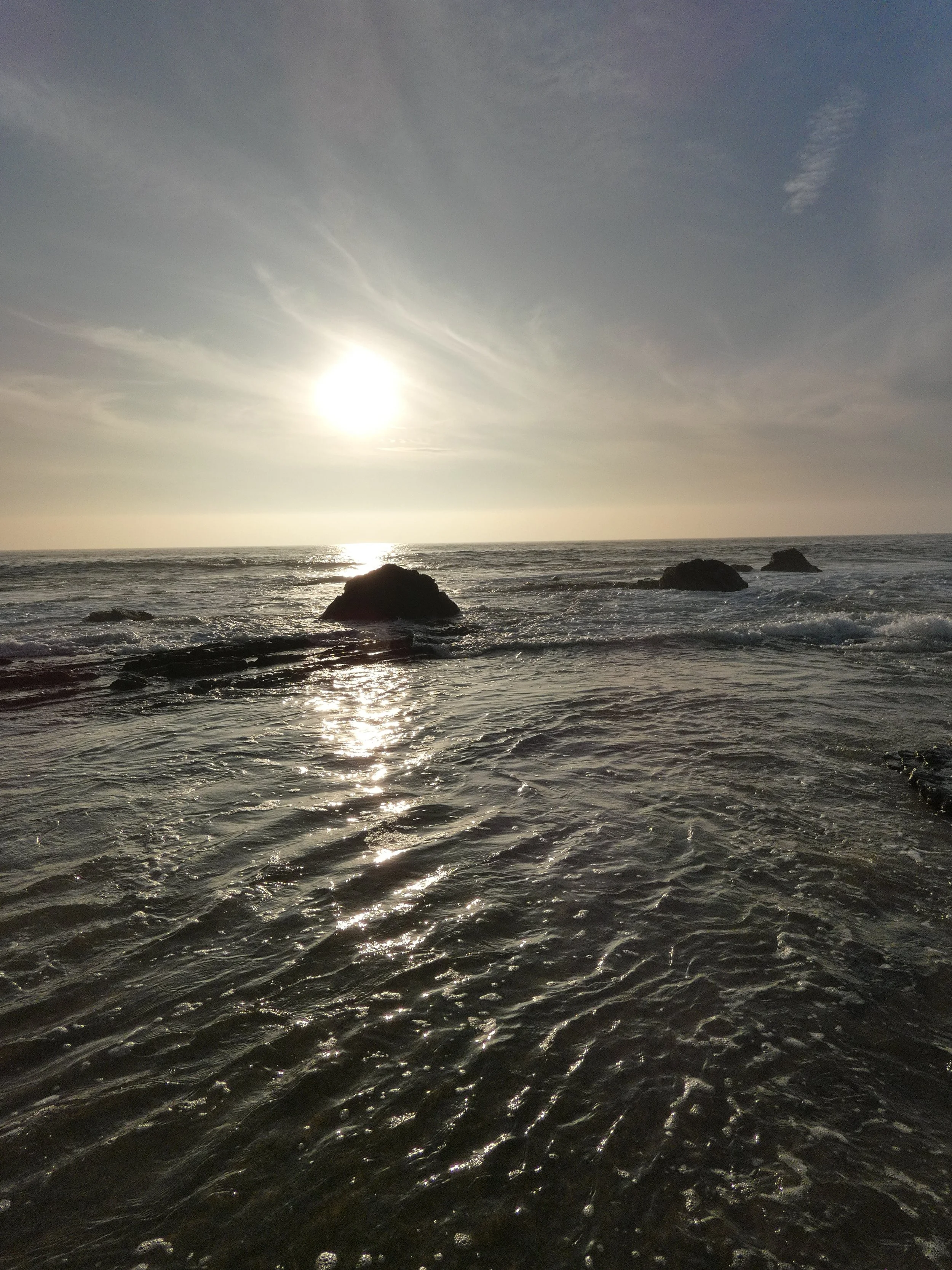 The image shows the ocean with three prominent rocks in the foreground and the sun shining brightly in the sky, reflecting off the water's surface, with wispy clouds in the background.