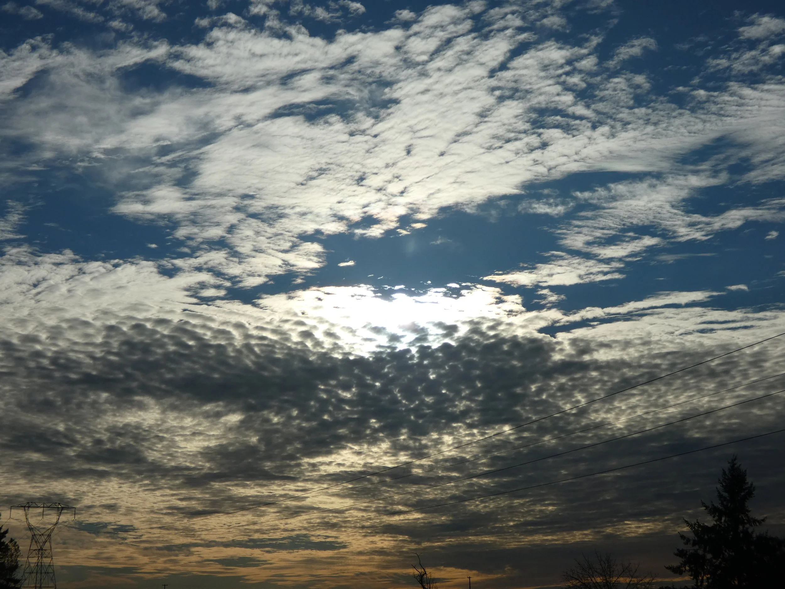 Cloudy sky with sunlight peeking through, power lines, and trees silhouetted along the horizon.
