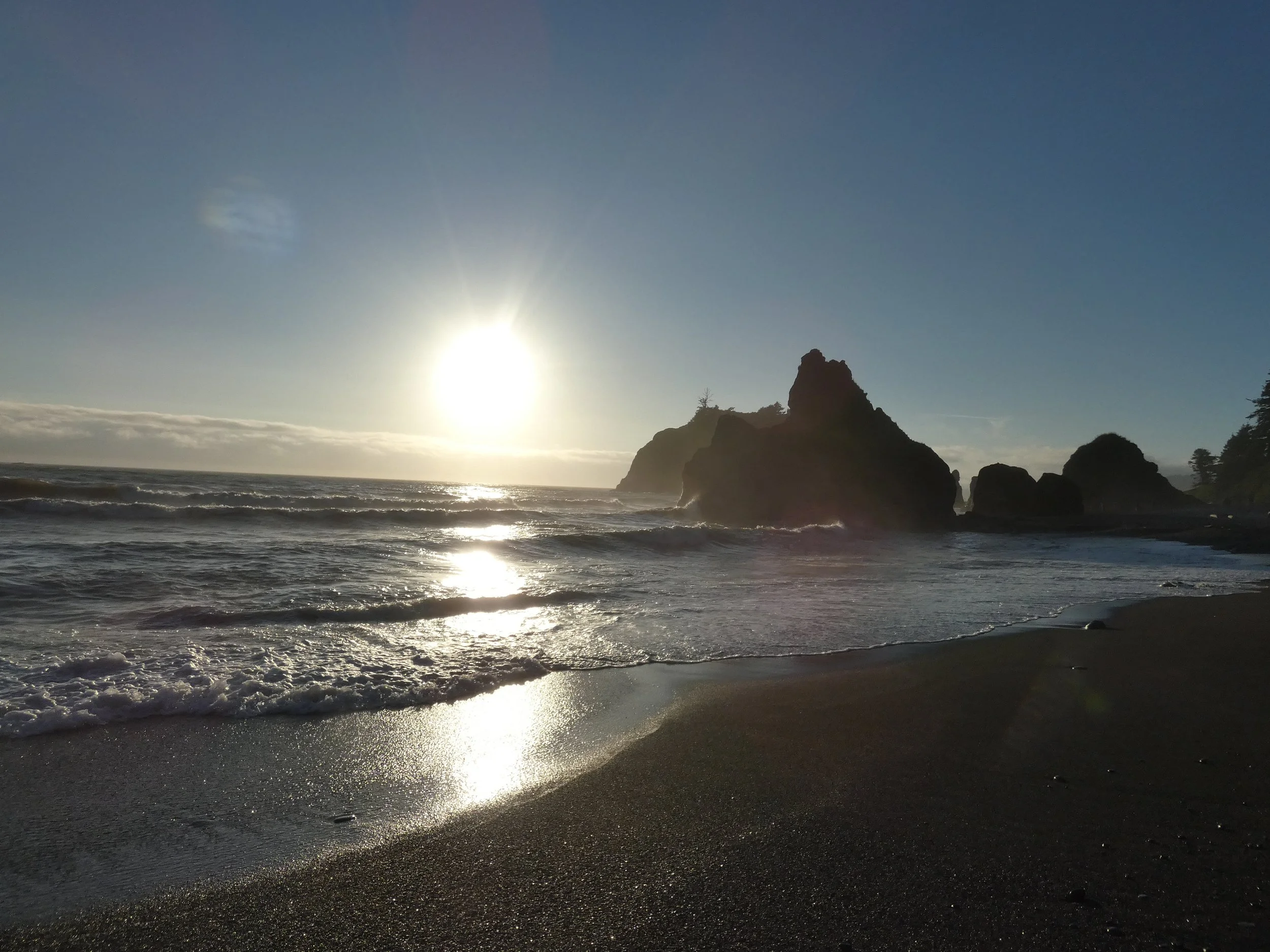 Sunset over the ocean with waves crashing onto the shore and large rocks along the coastline.