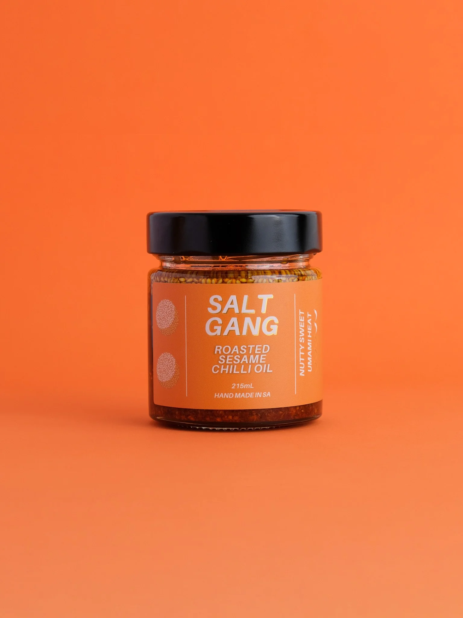 A jar of Salt Gang roasted sesame chili oil with a black lid, placed on an orange background. The label is orange with white text and mentions the flavor, volume, and country of origin.