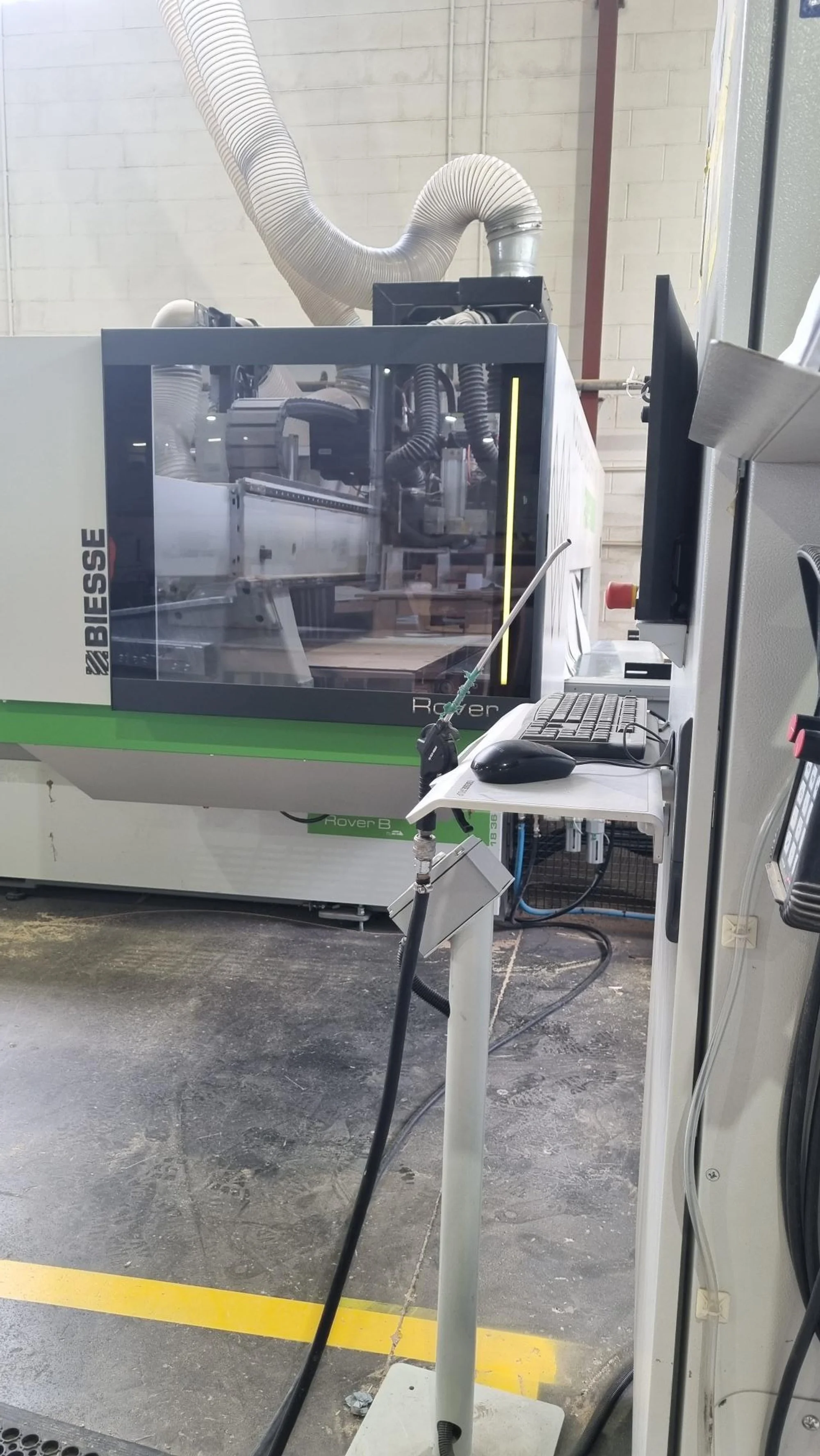 A Biesse Rover B CNC machine in a woodworking shop, with a computer monitor and mouse on a stand nearby, and a large flexible vent hose above to extract sawdust.