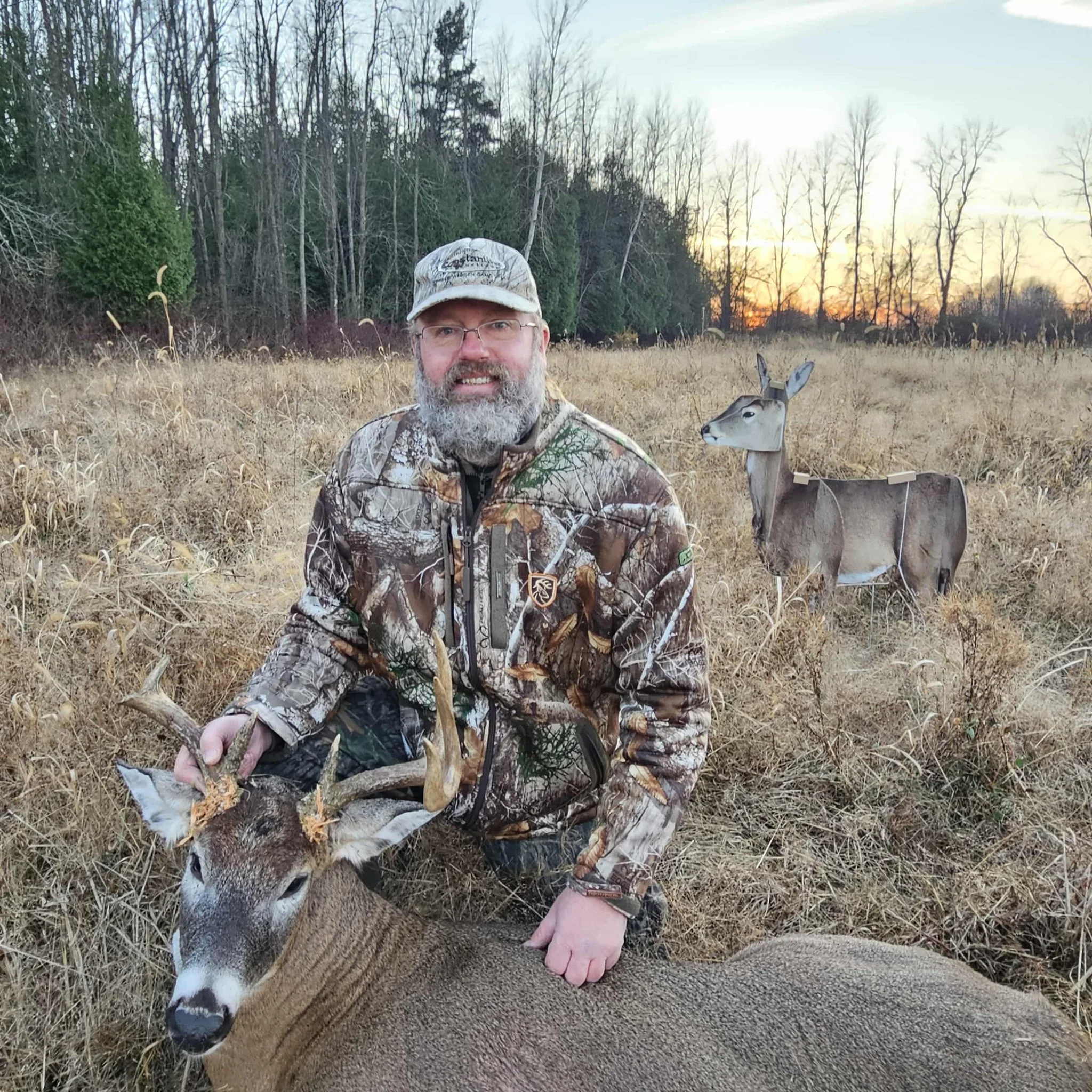 A man in camouflage clothing kneels in a field, holding the antlers of a harvested deer. Behind him, a cardboard cutout of a deer stands in the tall, dry grass as the sun sets in the background, with trees at the horizon.