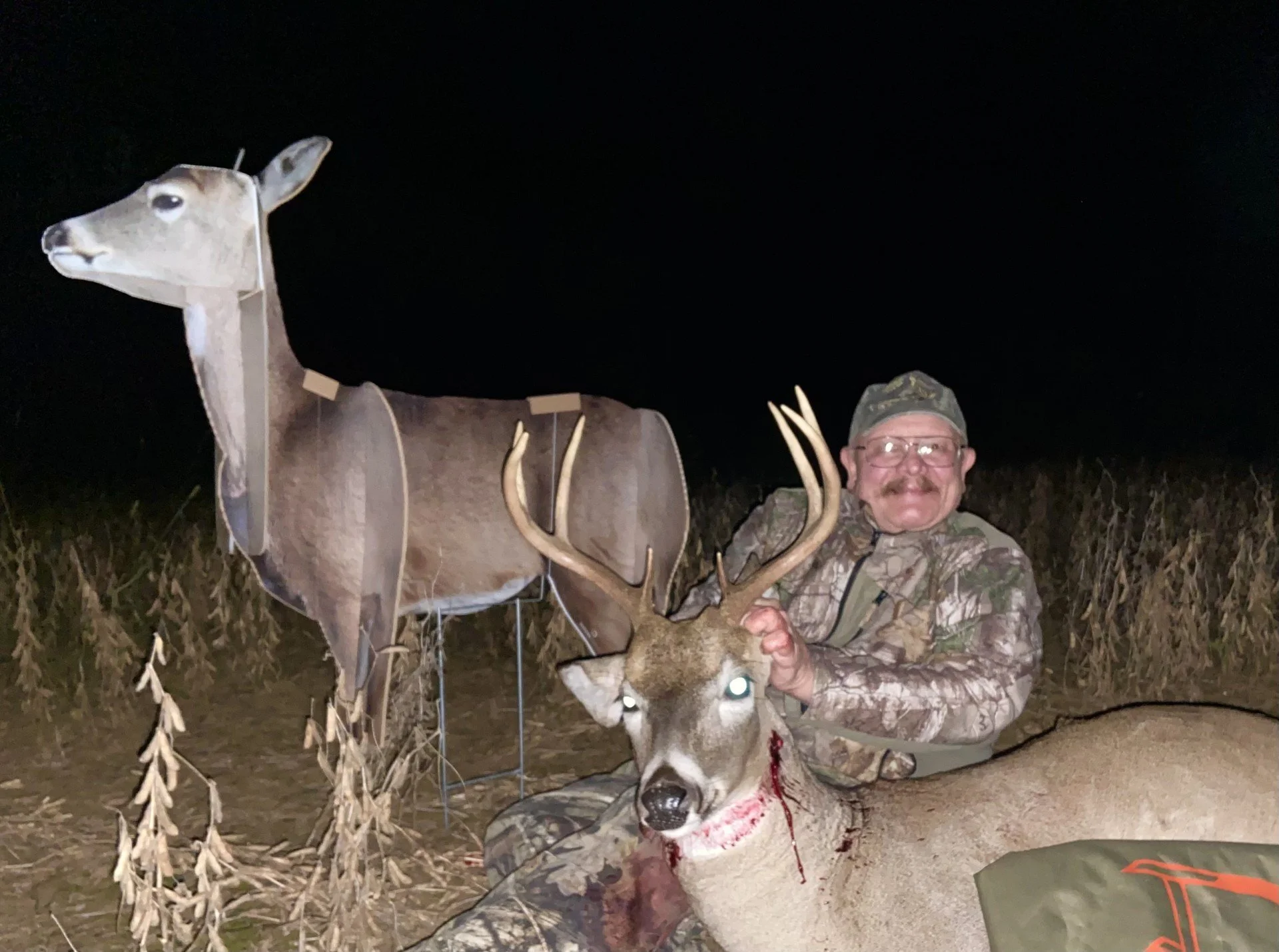 A man in camouflage clothing kneels with a large deer he has hunted, holding its antlers. The deer has a blood wound and glowing eyes. In the background, a decoy of a female deer is set up, and the scene takes place outdoors at night with a dark sky.