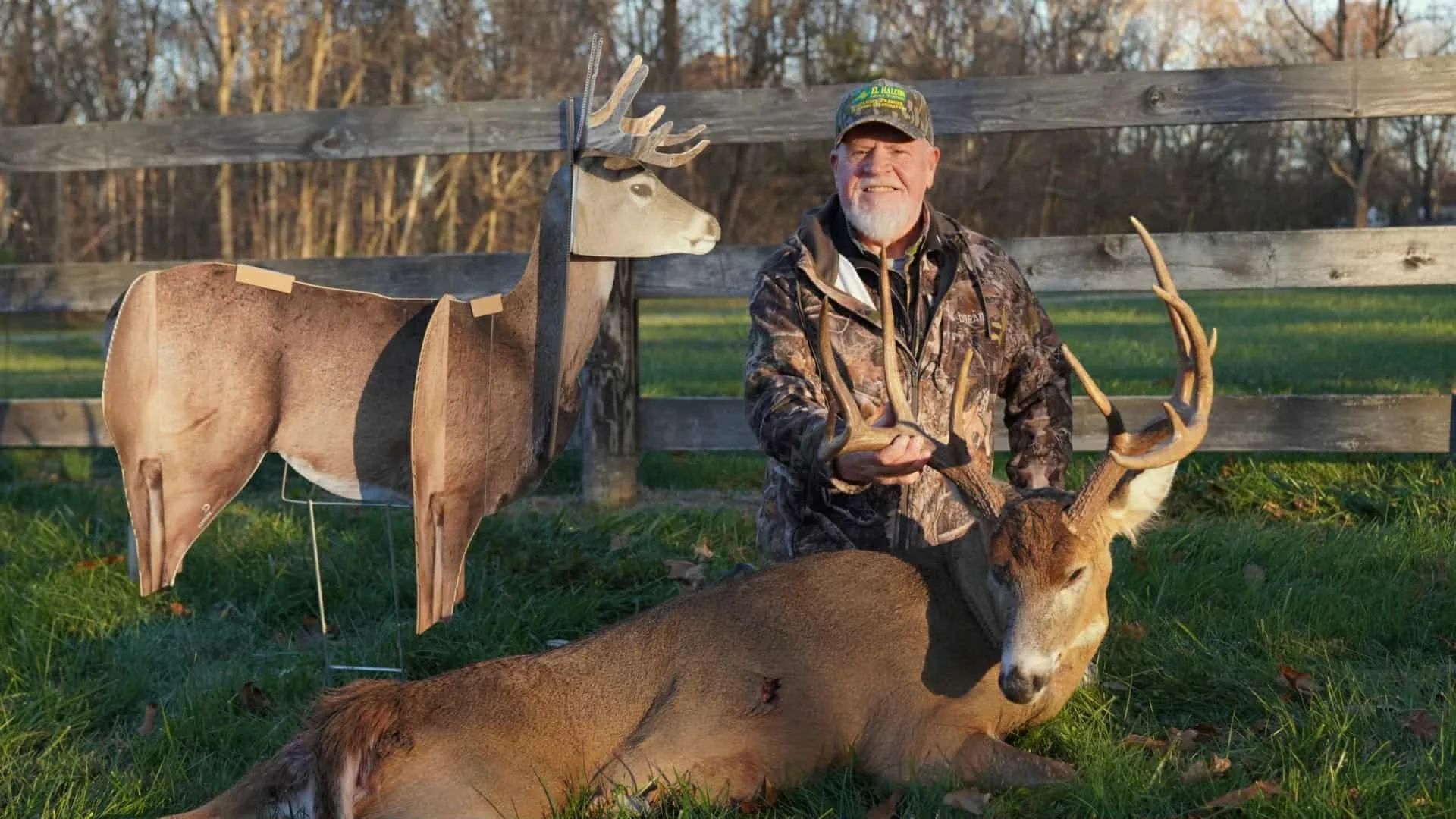 A man in camouflage clothing holding antlers of a large deer he hunts, sitting beside the dead deer laying on the grass with a paper deer cutout on a stand nearby, outdoor setting with a wooden fence and trees in the background.