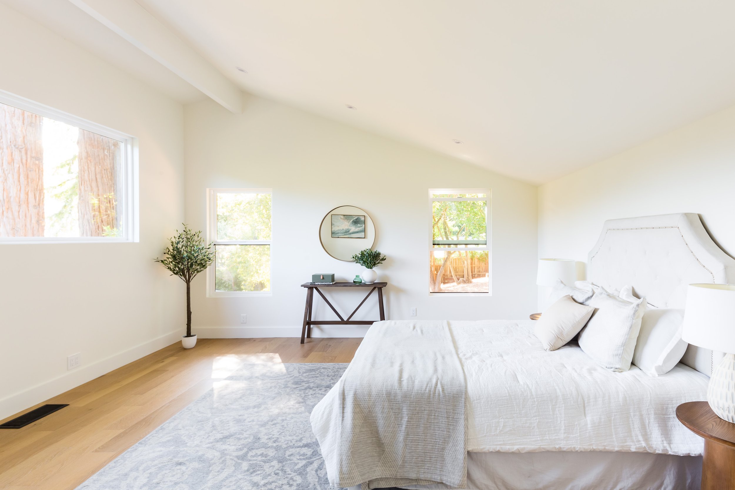 Bright and airy bedroom with light-colored bedding, a large white headboard, wooden side tables with lamps, a tall potted plant, and three windows bringing in natural light.