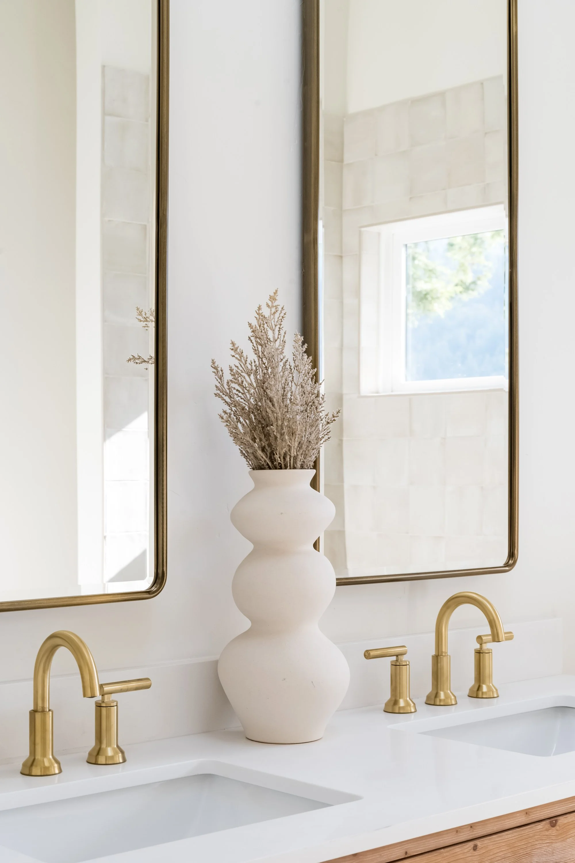 A minimalist bathroom sink area with a white countertop, dual sinks, gold faucets, a large decorative white vase with dried branches, two large gold-framed mirrors, and a window reflected in the mirrors.