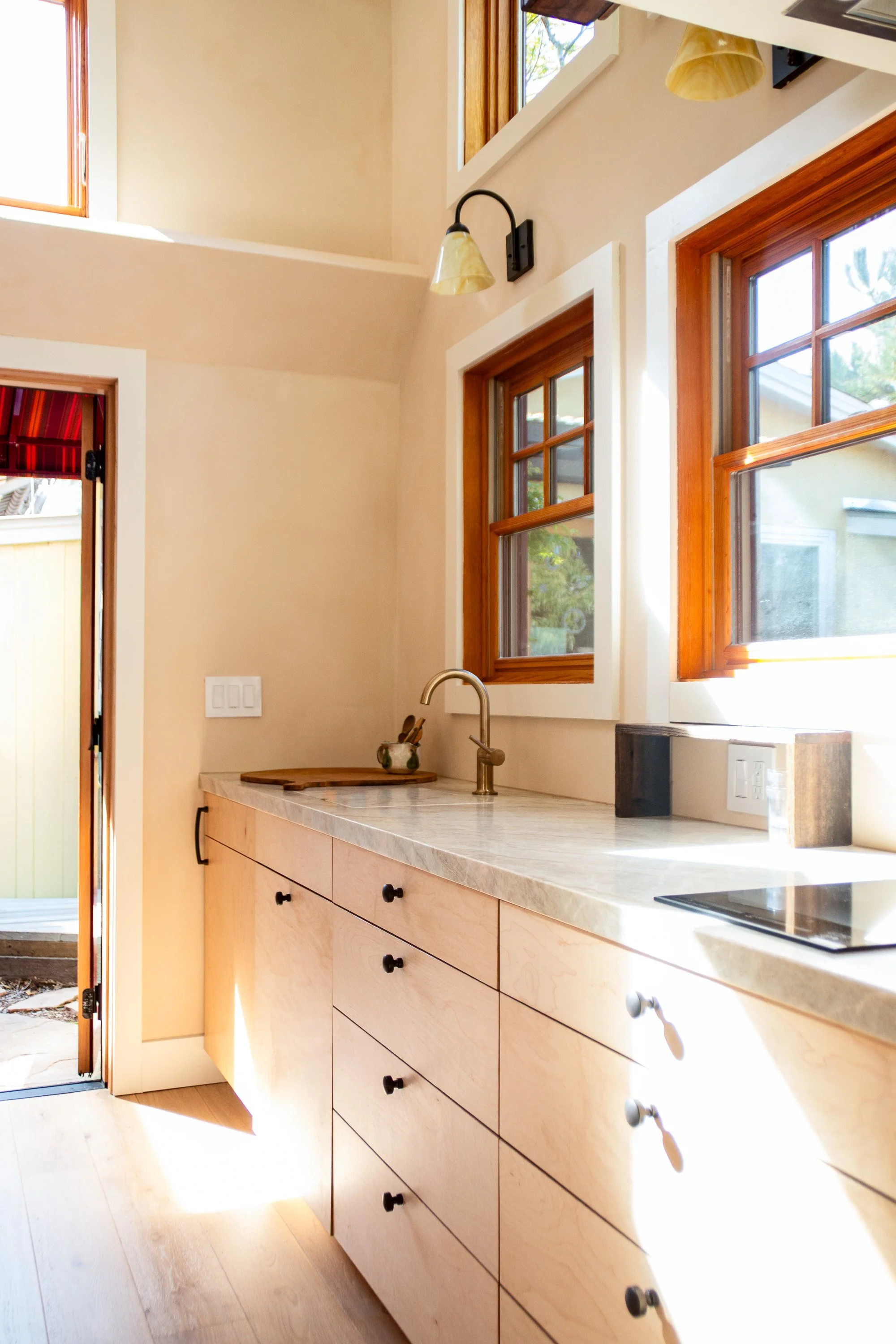 Bright kitchen with light wood cabinets, a marble countertop, two windows with wood frames, and a door leading outside. Natural sunlight illuminates the space.