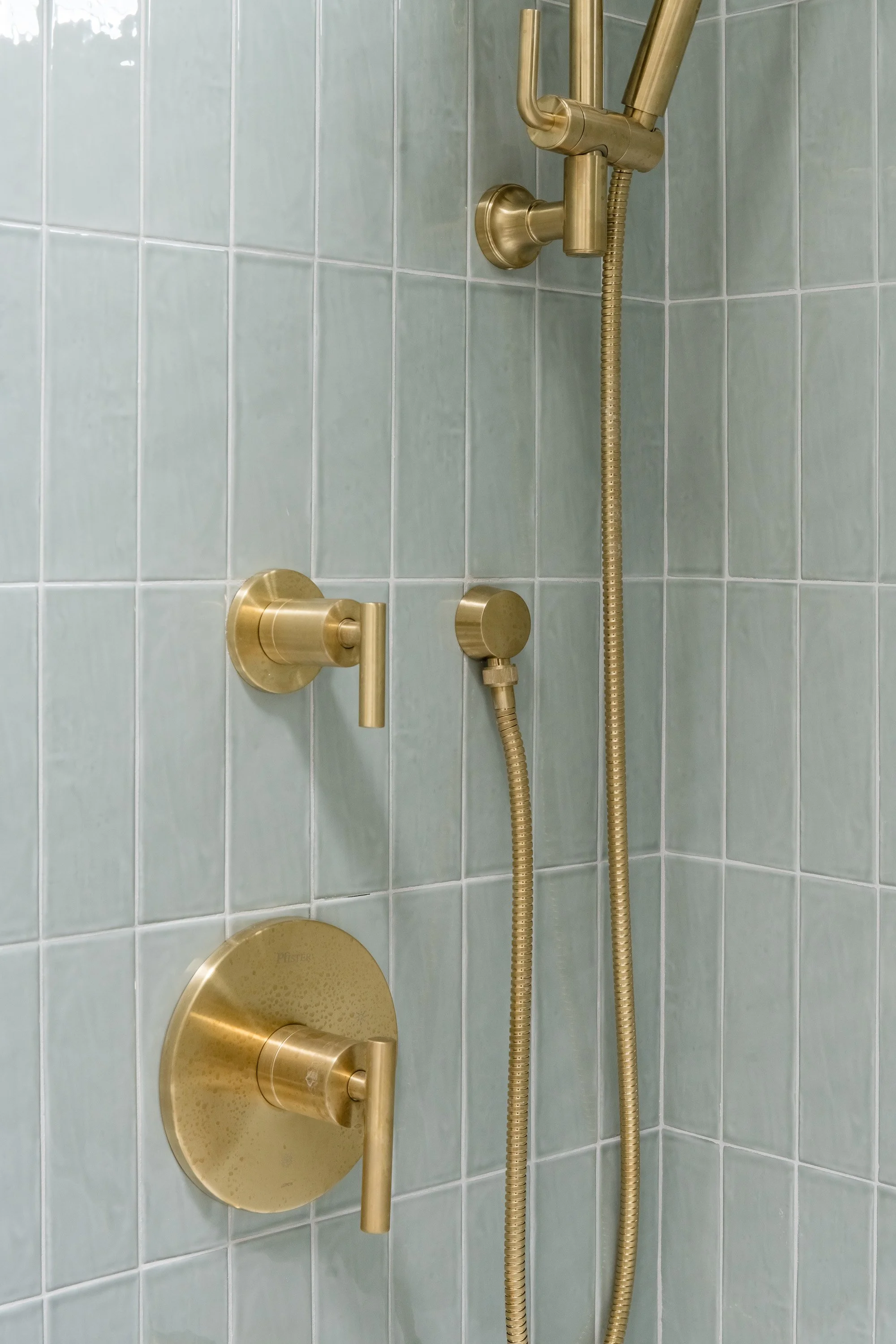 Shower fixtures including a brass showerhead, two brass handles, a brass control knob, and a brass handheld shower with a hose, mounted on a tiled wall in a bathroom.
