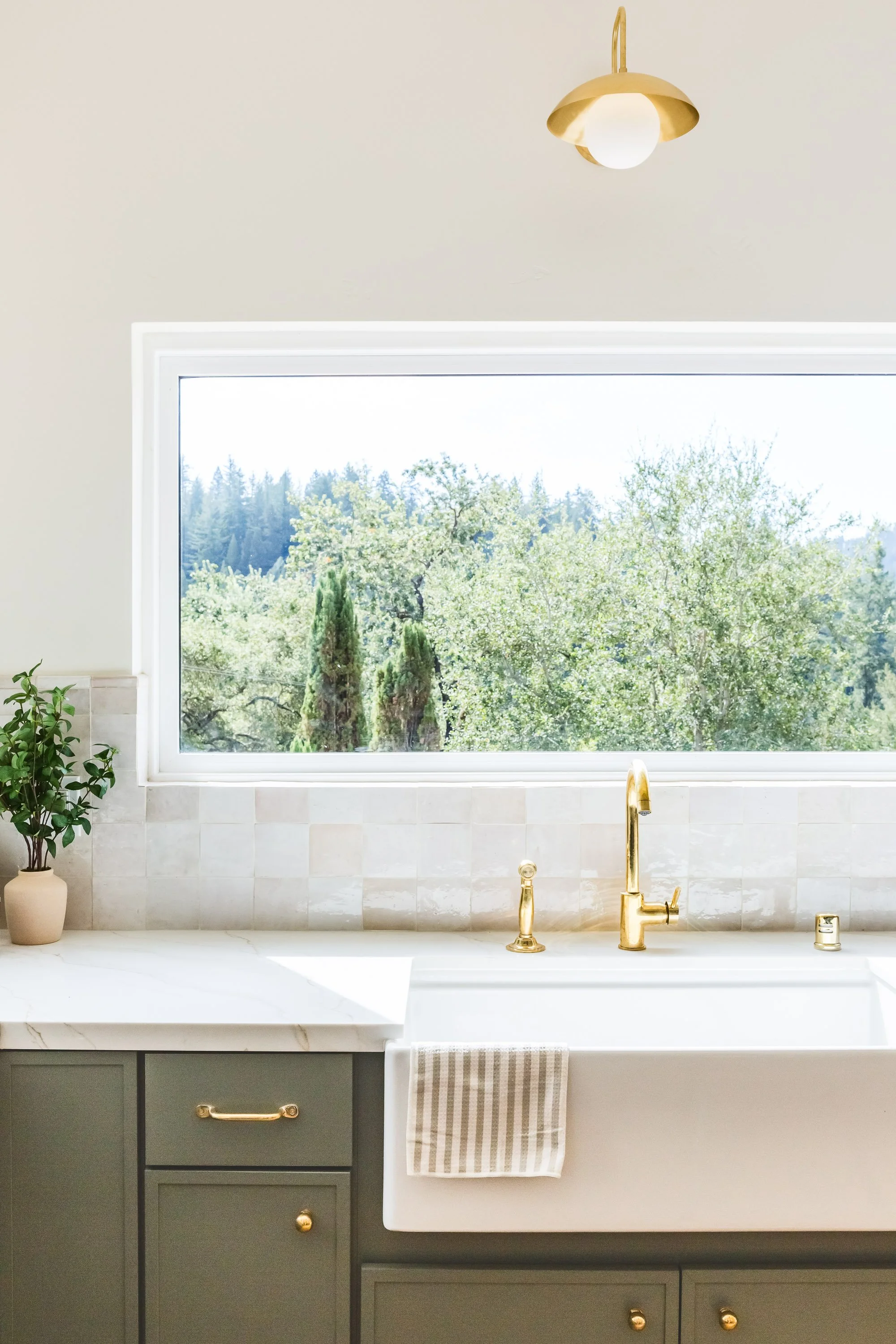 Bright kitchen with a large window overlooking trees, gold faucet and fixtures, green cabinet, potted plant, and striped towel.