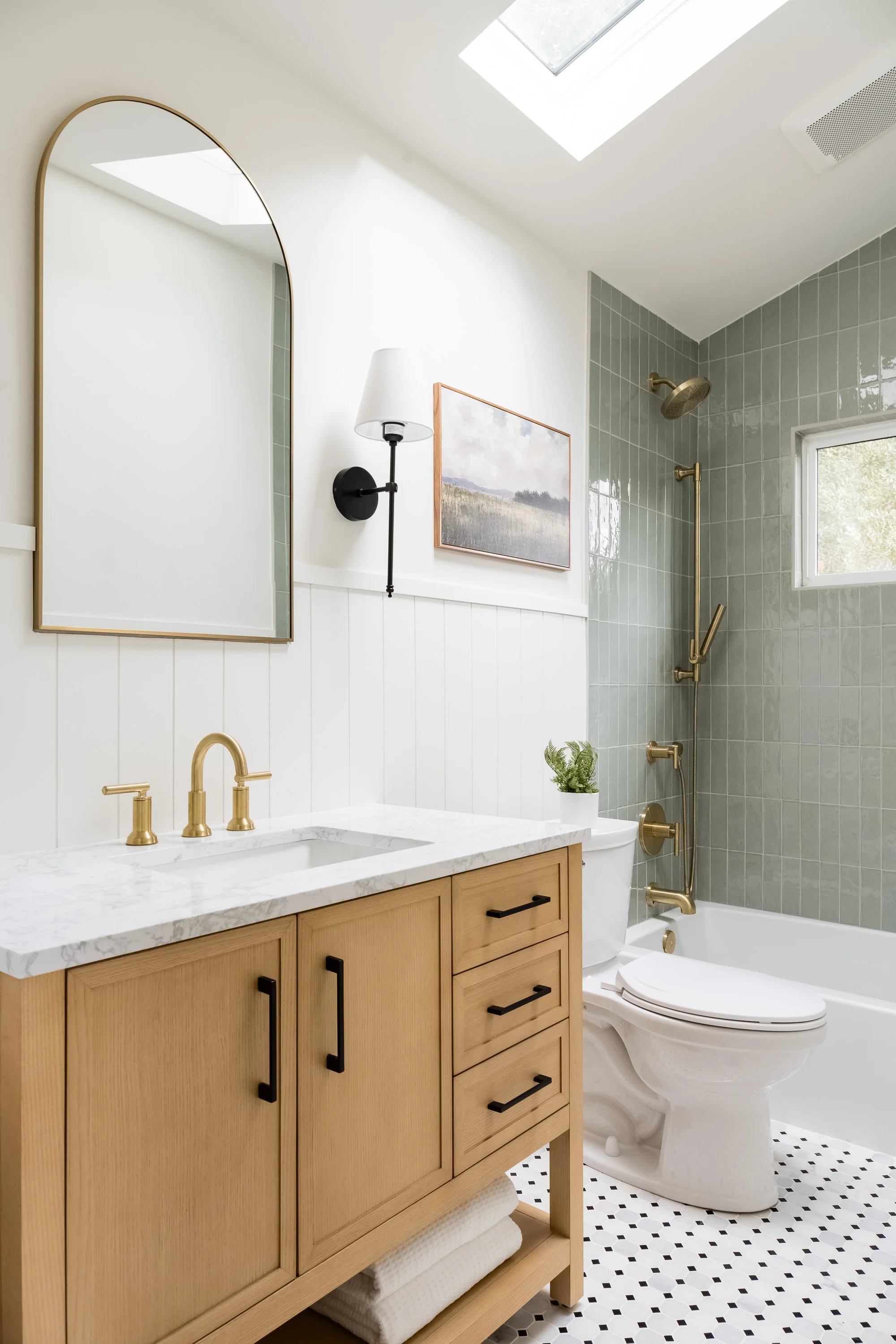 Modern bathroom with wooden cabinet, marble countertop, gold fixtures, white toilet, bathtub with gold shower fixtures, small window, skylight, black and white tile floor, wall art, mirror, and wall-mounted light.