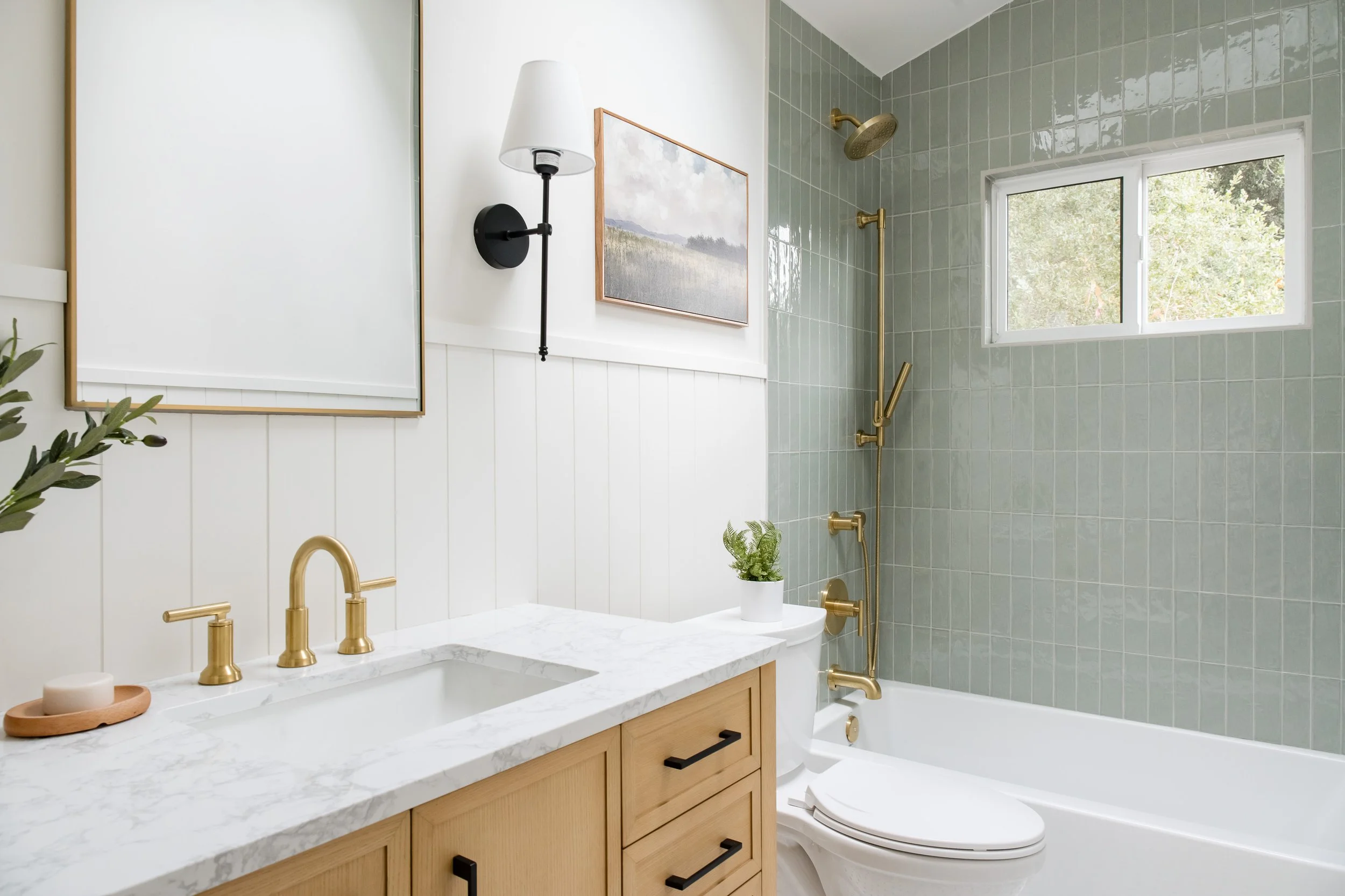 Bathroom with a white marble vanity, gold fixtures, a bathtub with a gold shower head, green tiled wall, window, and framed artwork