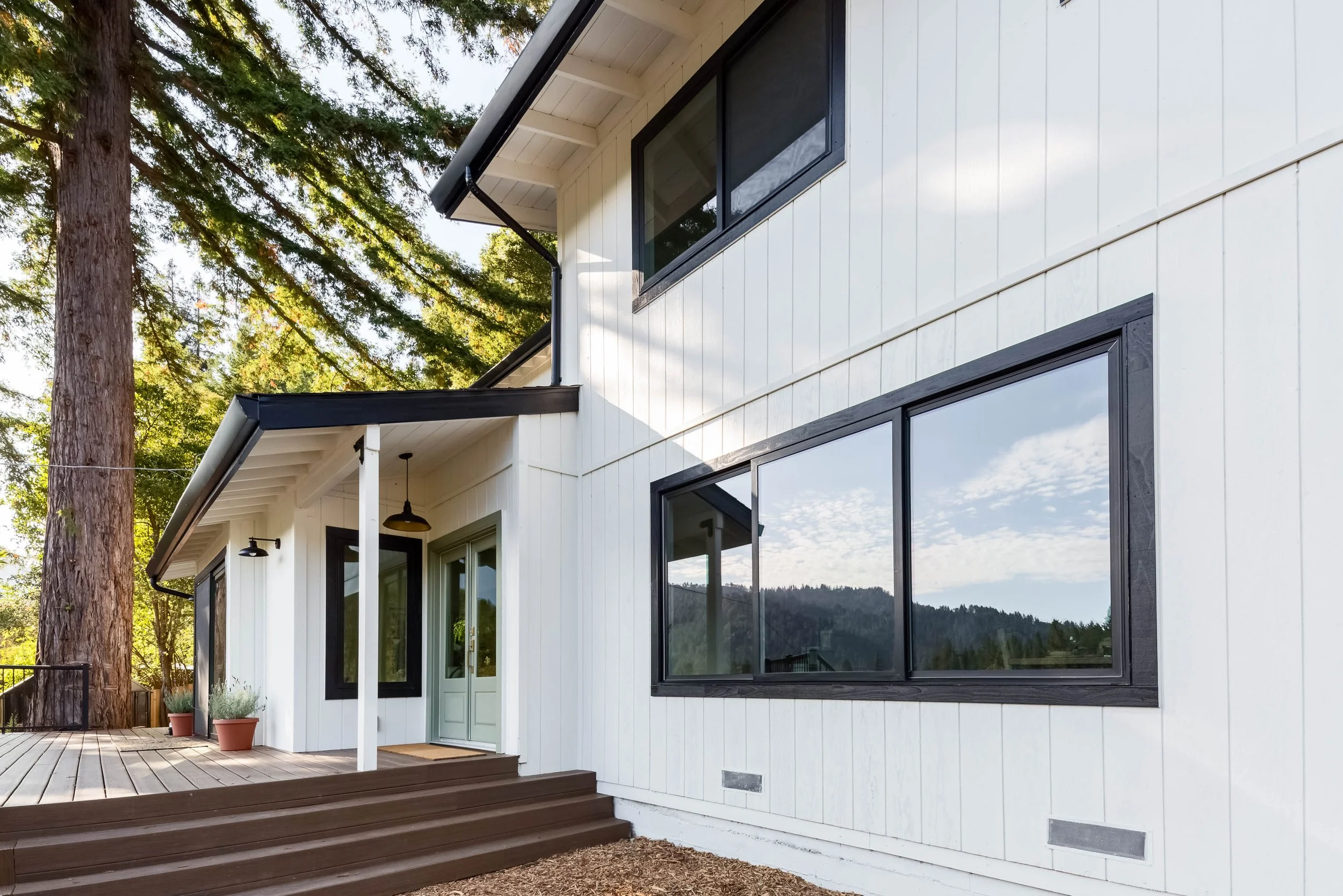 Modern white house with large windows, black framing, and a small porch with steps, surrounded by tall trees, in a natural landscape.