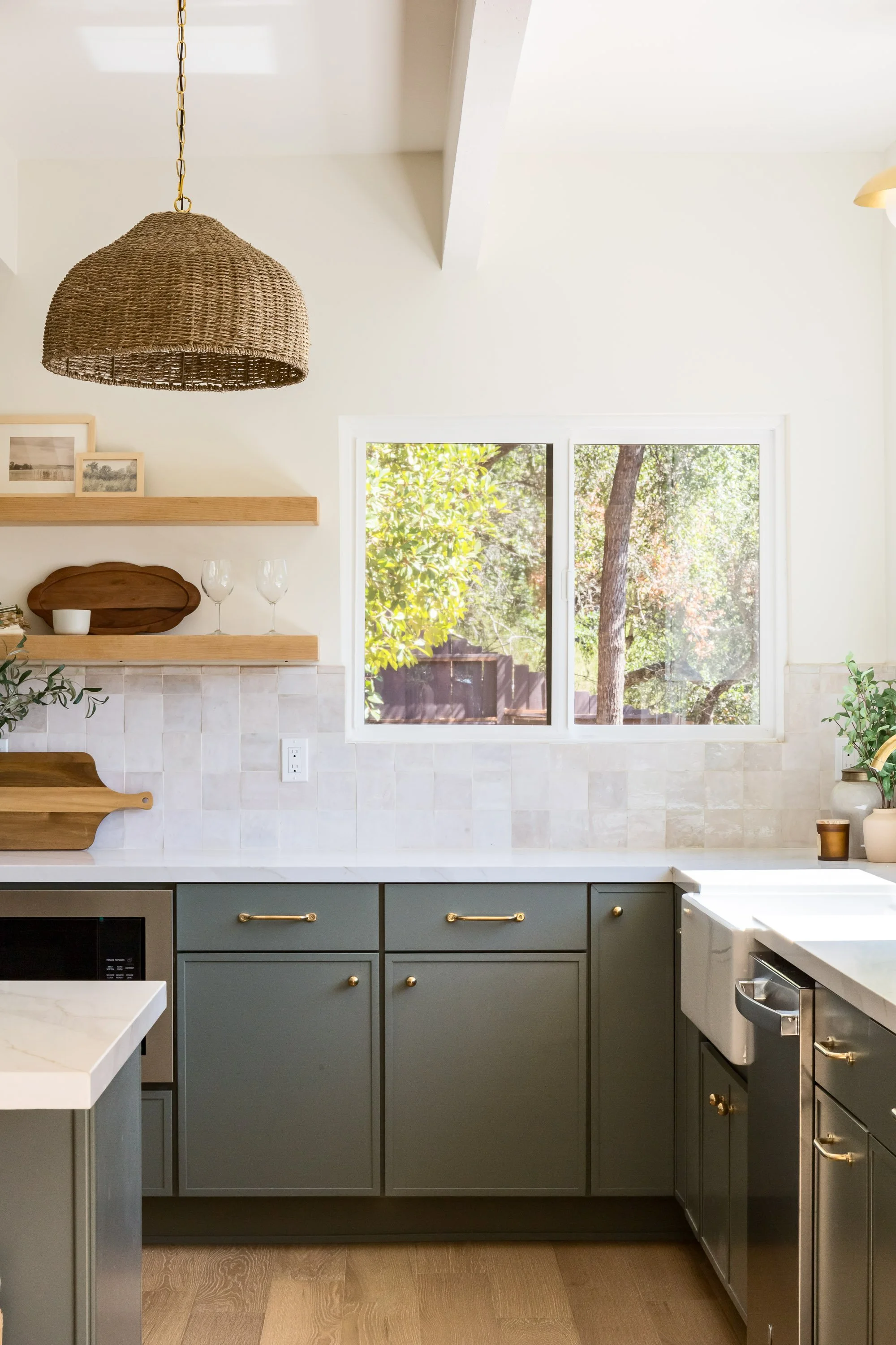 A kitchen with gray cabinets, white countertops, a window showing trees outside, and decorative shelves with framed photos and wine glasses.