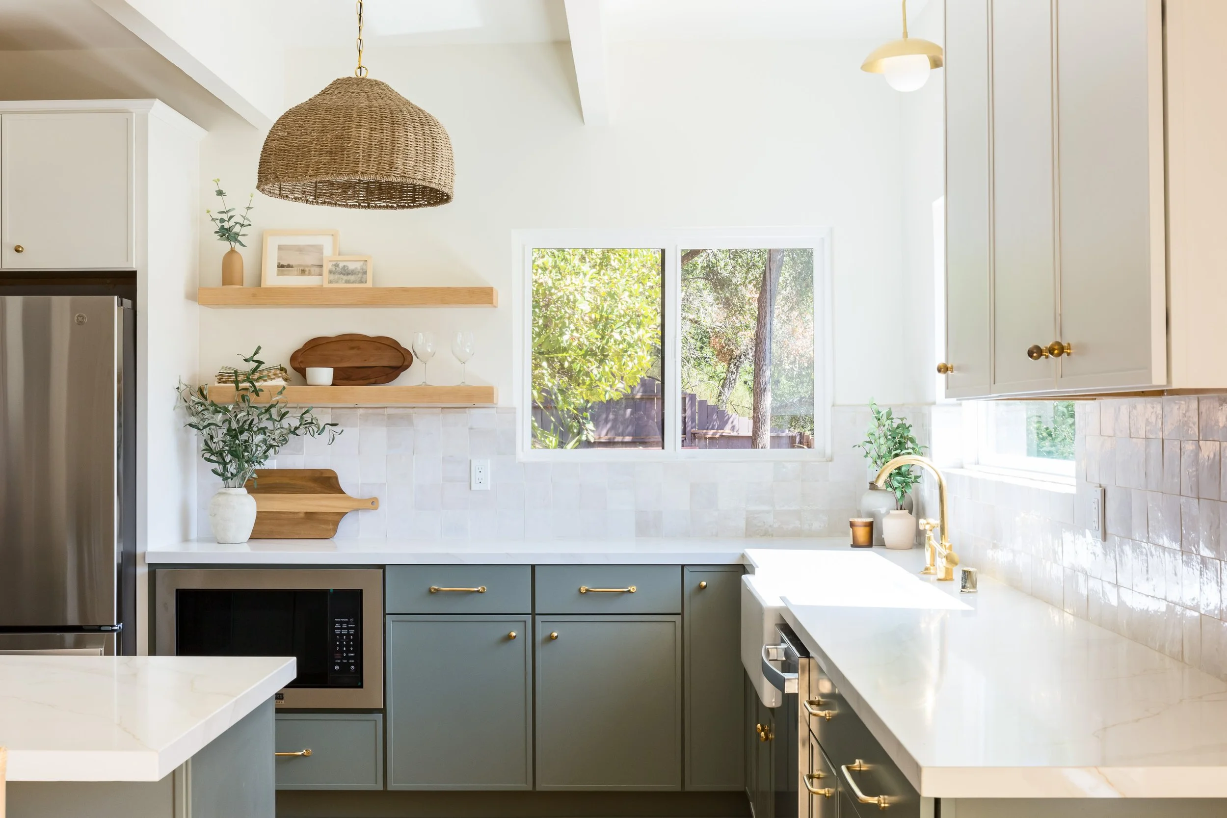 Bright kitchen with light gray lower cabinets, white upper cabinets, white countertop, and a window overlooking a backyard with trees. Shelves above the counter display framed photos, a vase with greenery, a wooden tray, and wine glasses. A woven pen