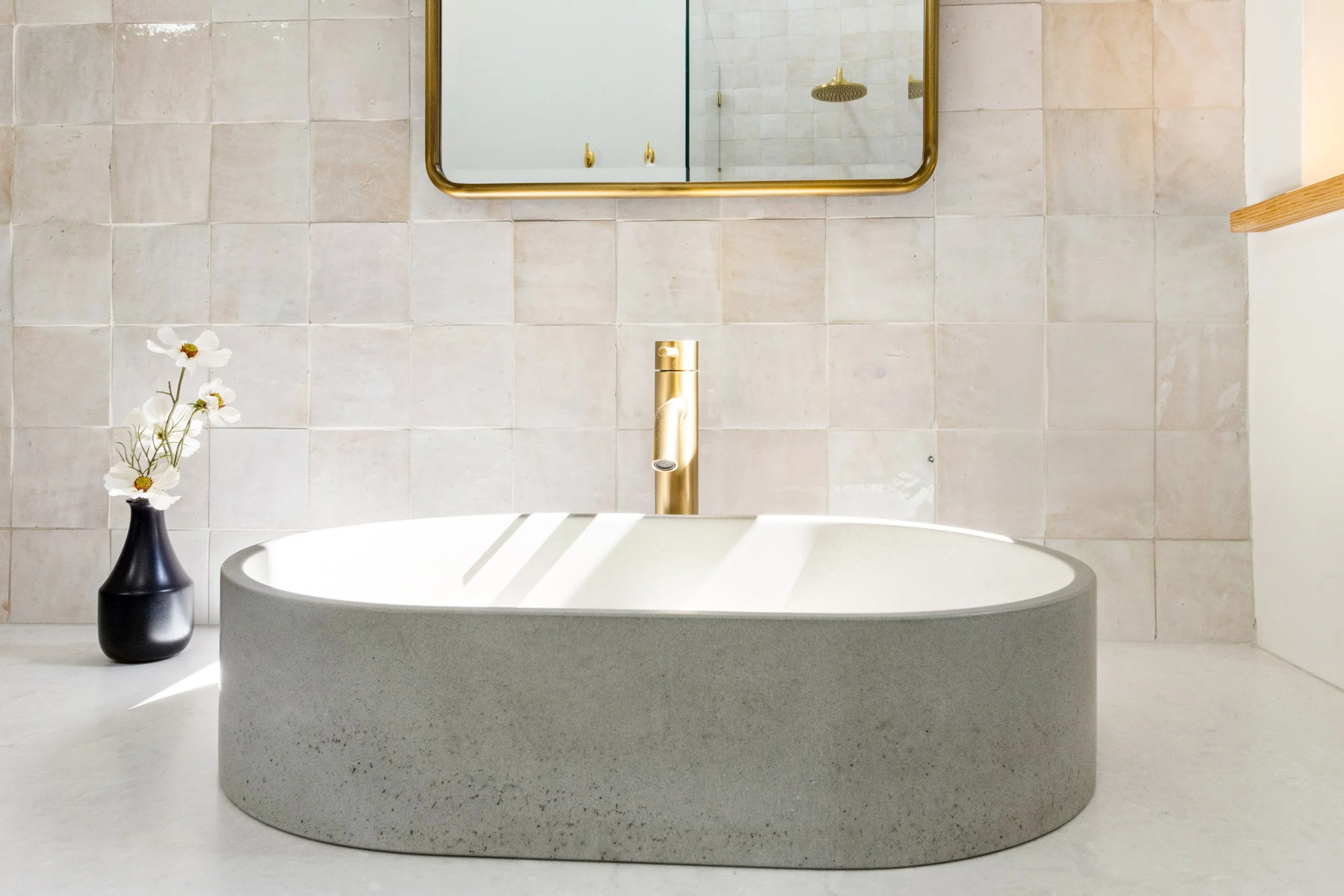 Modern bathroom with a large white bathtub, gold faucet, and a black vase with white flowers on the left.