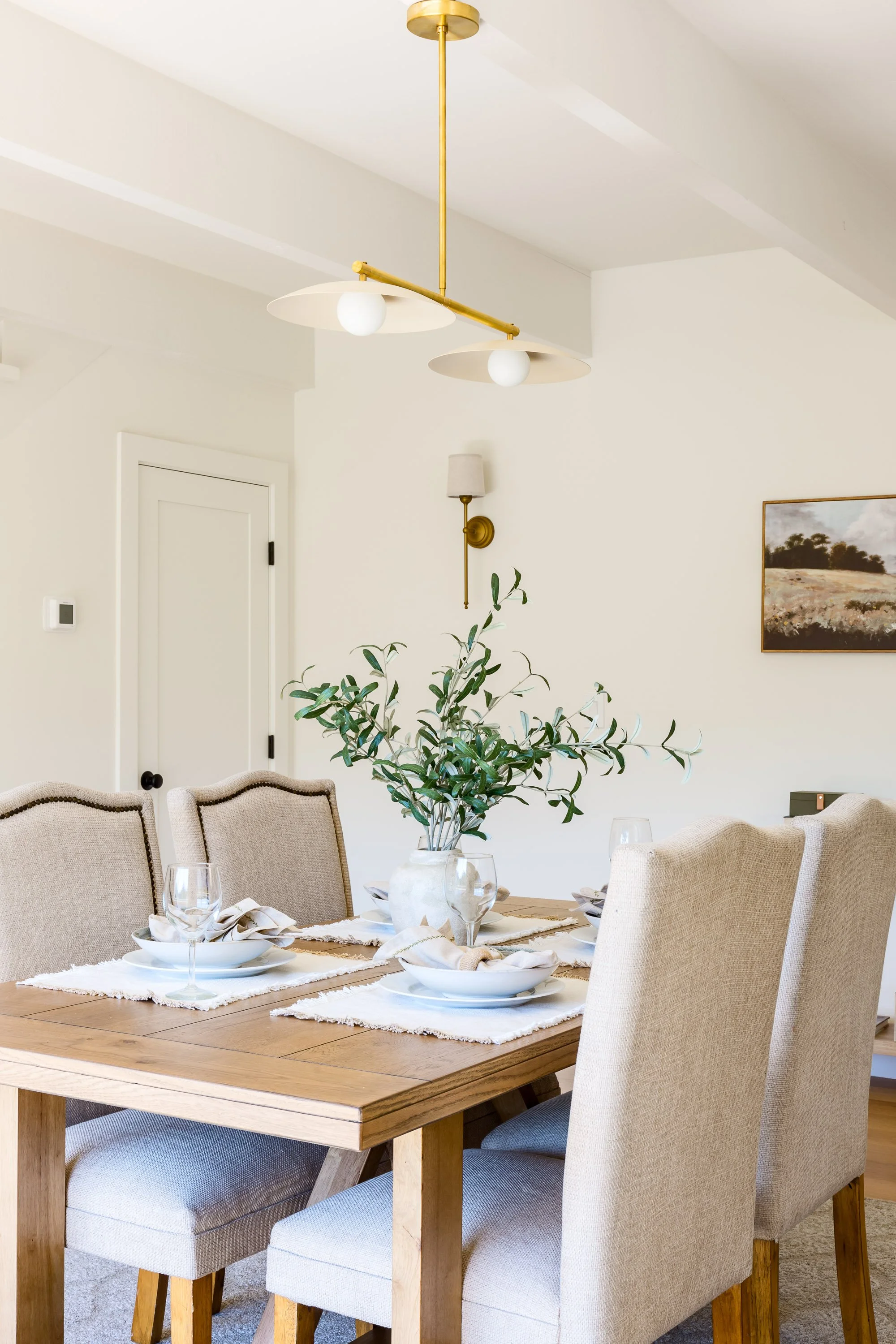 A dining room with a wooden table set for four, featuring white plates, glasses, and beige napkins. A large potted plant is in the center. The room has beige upholstered chairs, a ceiling light fixture with two white shades, a wall sconce, and a fram