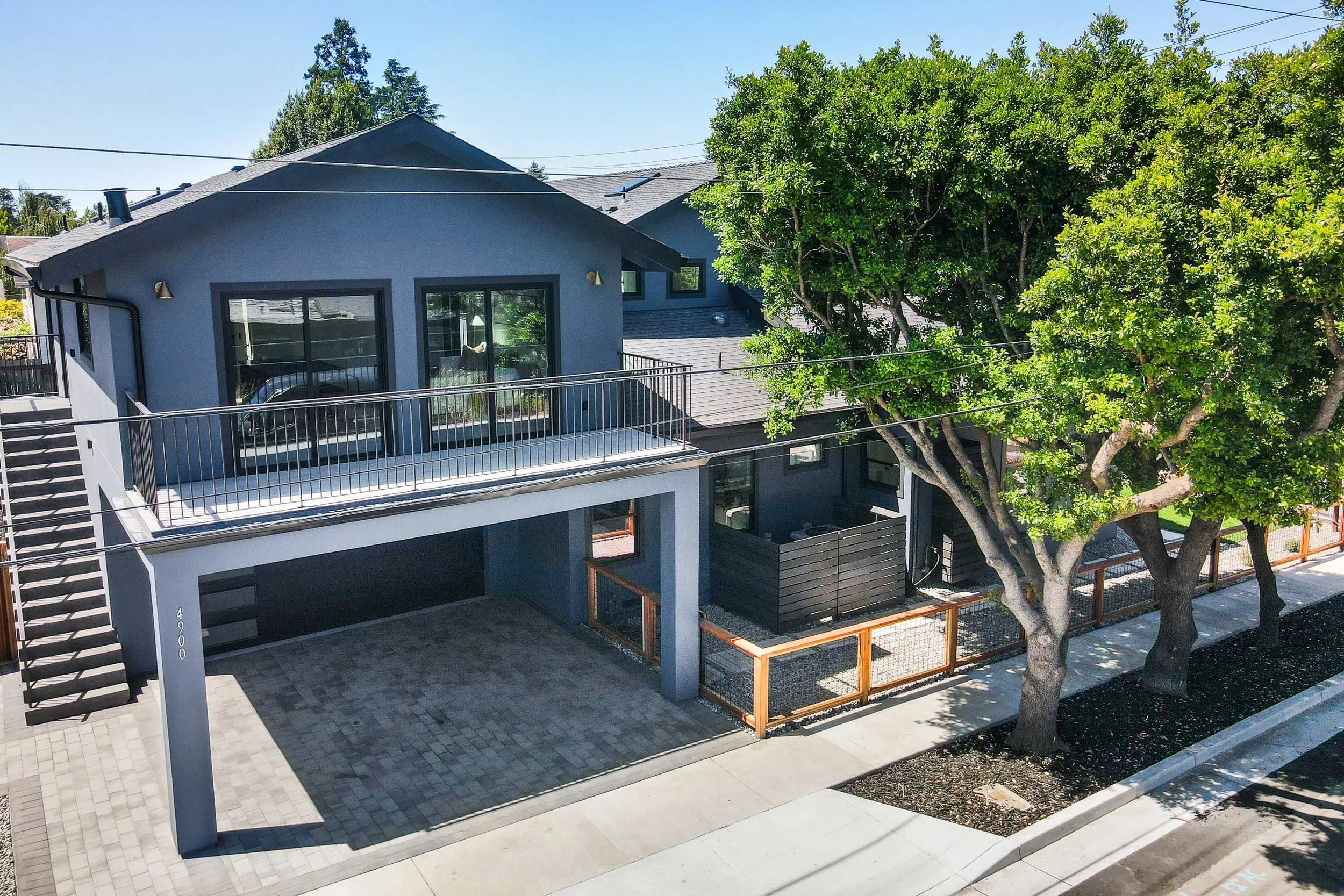 Modern two-story house with a rooftop deck, large windows, gray exterior, surrounded by trees with a sidewalk in front and a paved driveway.