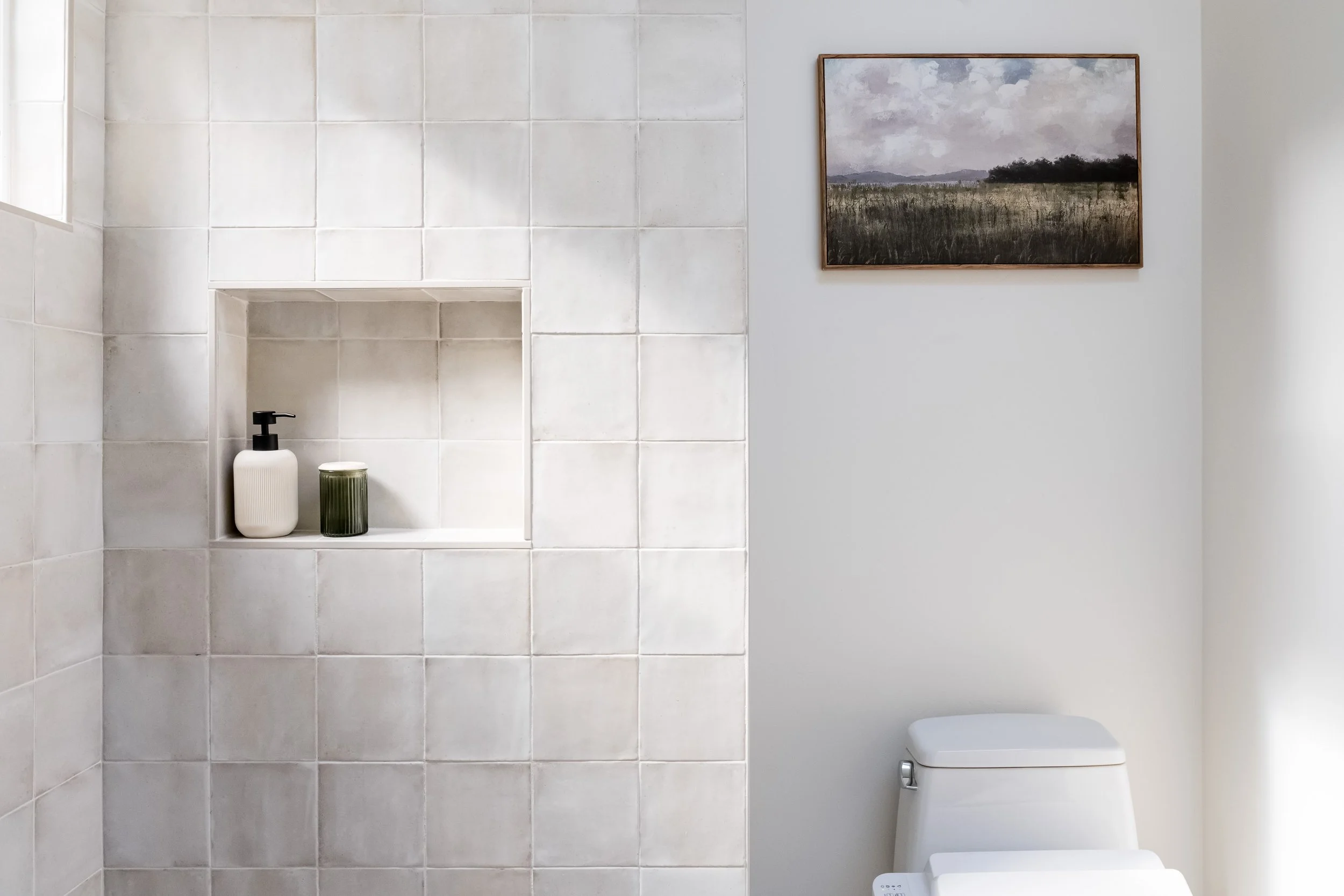 A modern bathroom with tiled shower wall, a white toilet, a built-in shower niche with soap dispenser and candle, and a landscape painting on the wall.