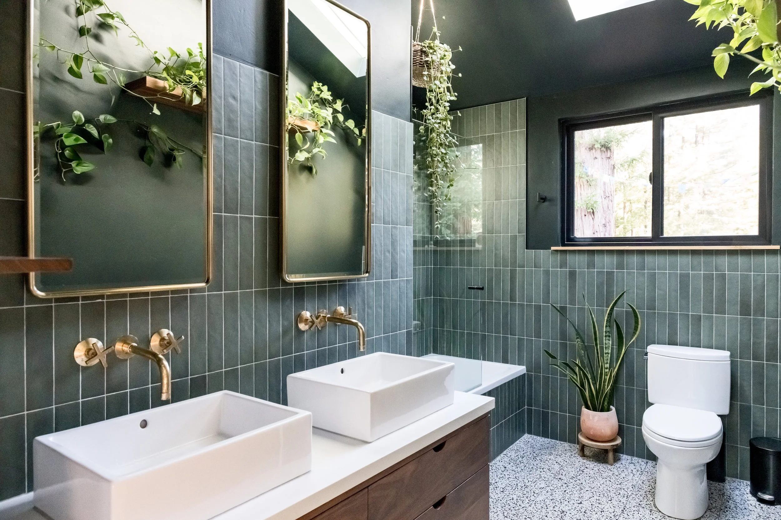 Modern bathroom with dark green tiled walls, double white rectangular sinks with gold faucets, a window, and green plants.