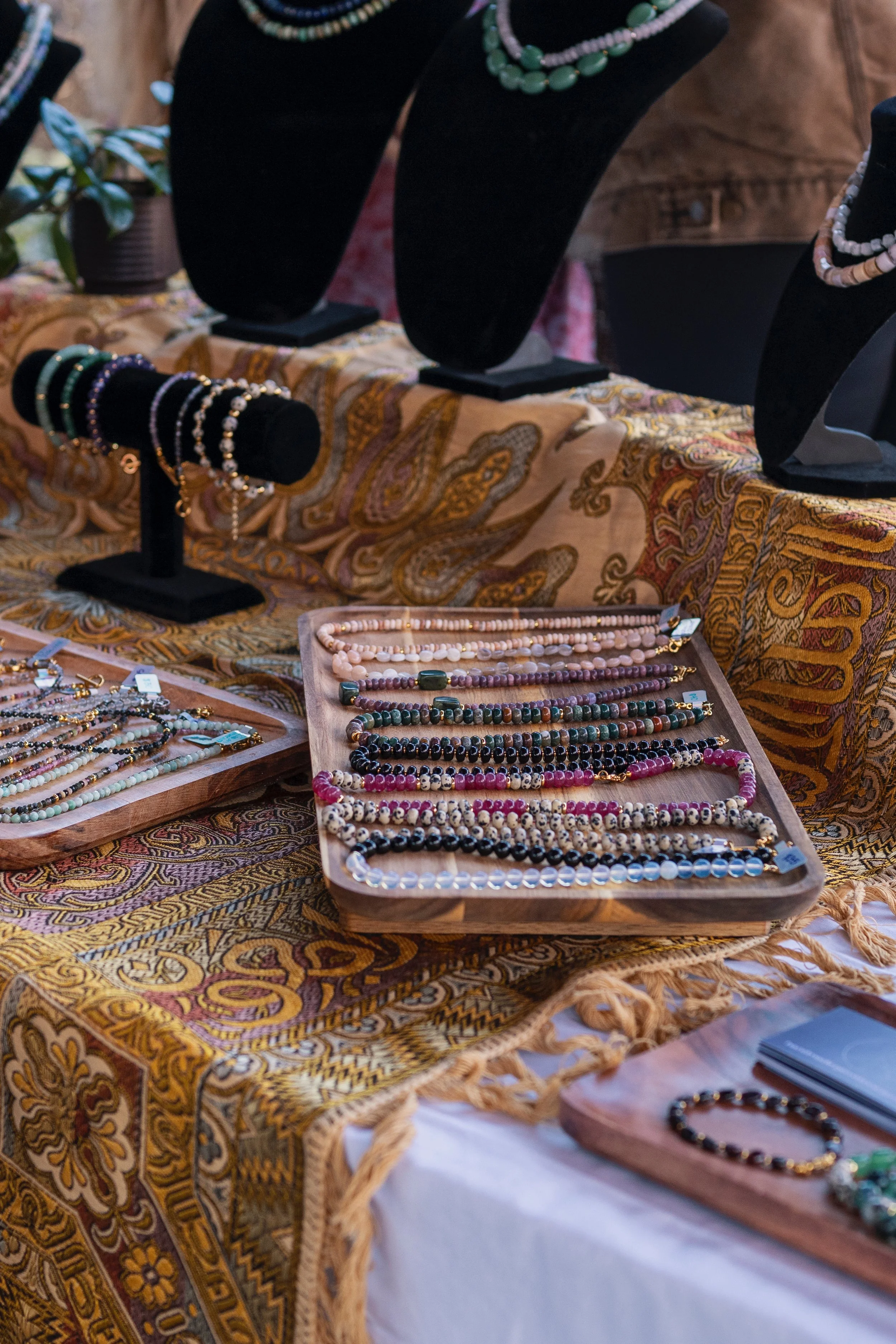 Jewelry display with various beaded necklaces and bracelets at a market stall, arranged on trays and busts, with a colorful patterned cloth.