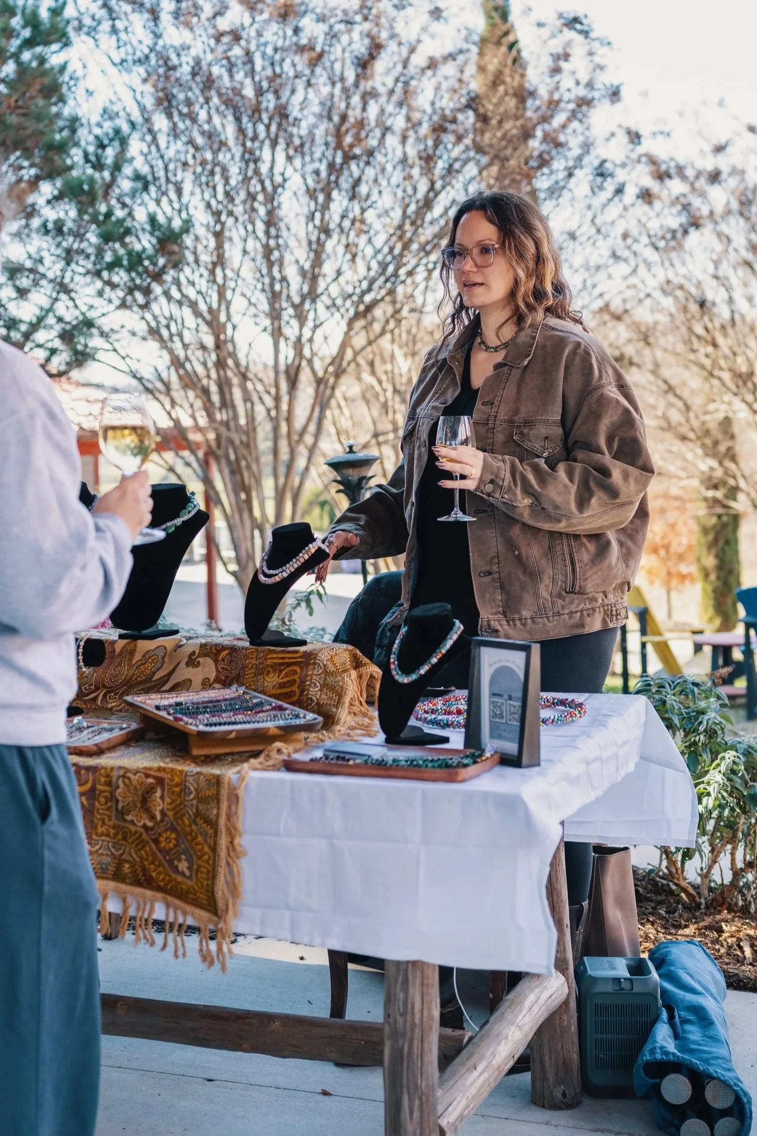 A woman at an outdoor jewelry booth holding a glass of wine, talking to a customer with a similar glass. The booth displays necklaces and jewelry on black busts and trays.