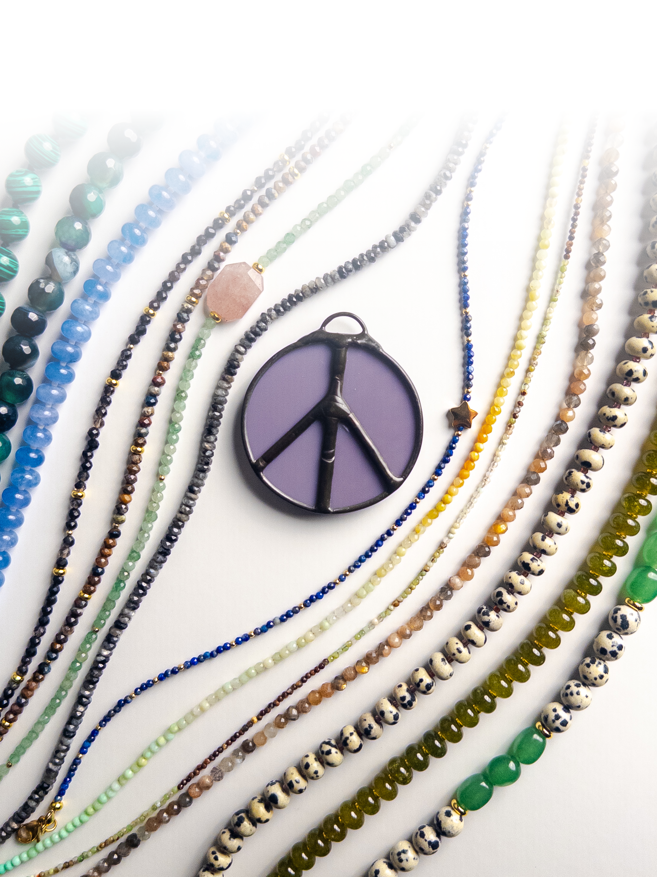 Colorful beaded necklaces arranged in parallel lines on a white surface with a lavender peace sign pendant in the center.