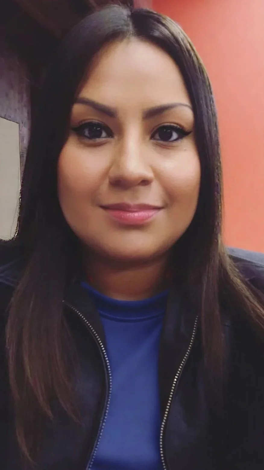 Close-up photo of a woman with straight, dark brown hair, wearing a blue top and a black jacket. She is smiling softly, with a warm, reddish background, and Spanish speaking immigrant consultant.