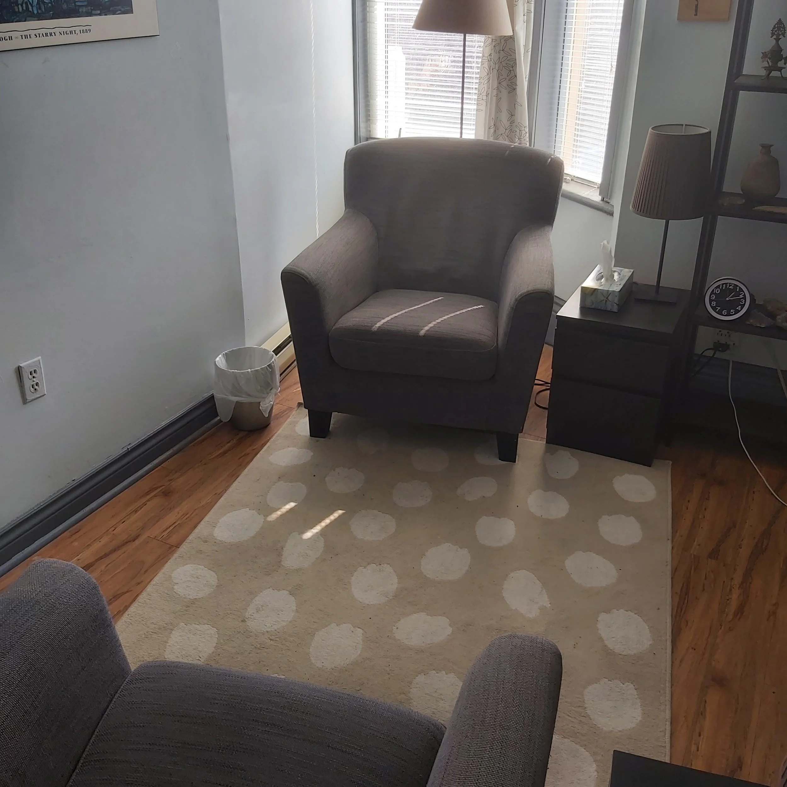 A cozy living room corner with a gray armchair placed on a beige rug with white polka dots. Next to the armchair is a black side table holding a tissue box and a lamp with a brown shade. A larger lamp with a beige shade is on the windowsill behind the armchair. The window has blinds and floral curtains. A framed picture hangs on the white wall to the left of the window, and an open black shelving unit displays decorative vases and a clock.