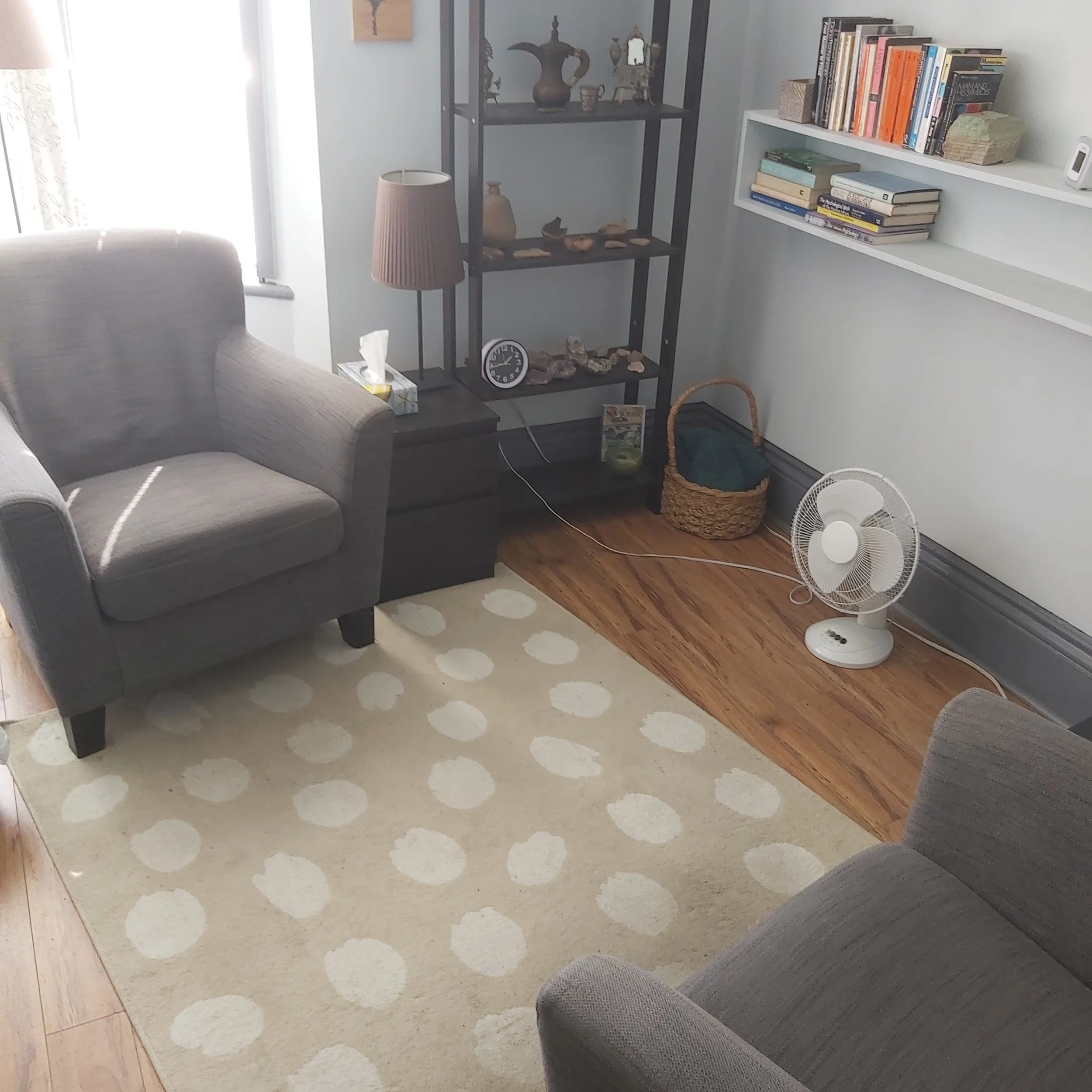 A cozy living room corner with a gray armchair, a black bookshelf decorated with pottery, a small black side table with a lamp and tissue box, a basket on the floor, a white oscillating fan, a beige polka dot rug, wooden flooring, and a wall-mounted white bookshelf filled with books.