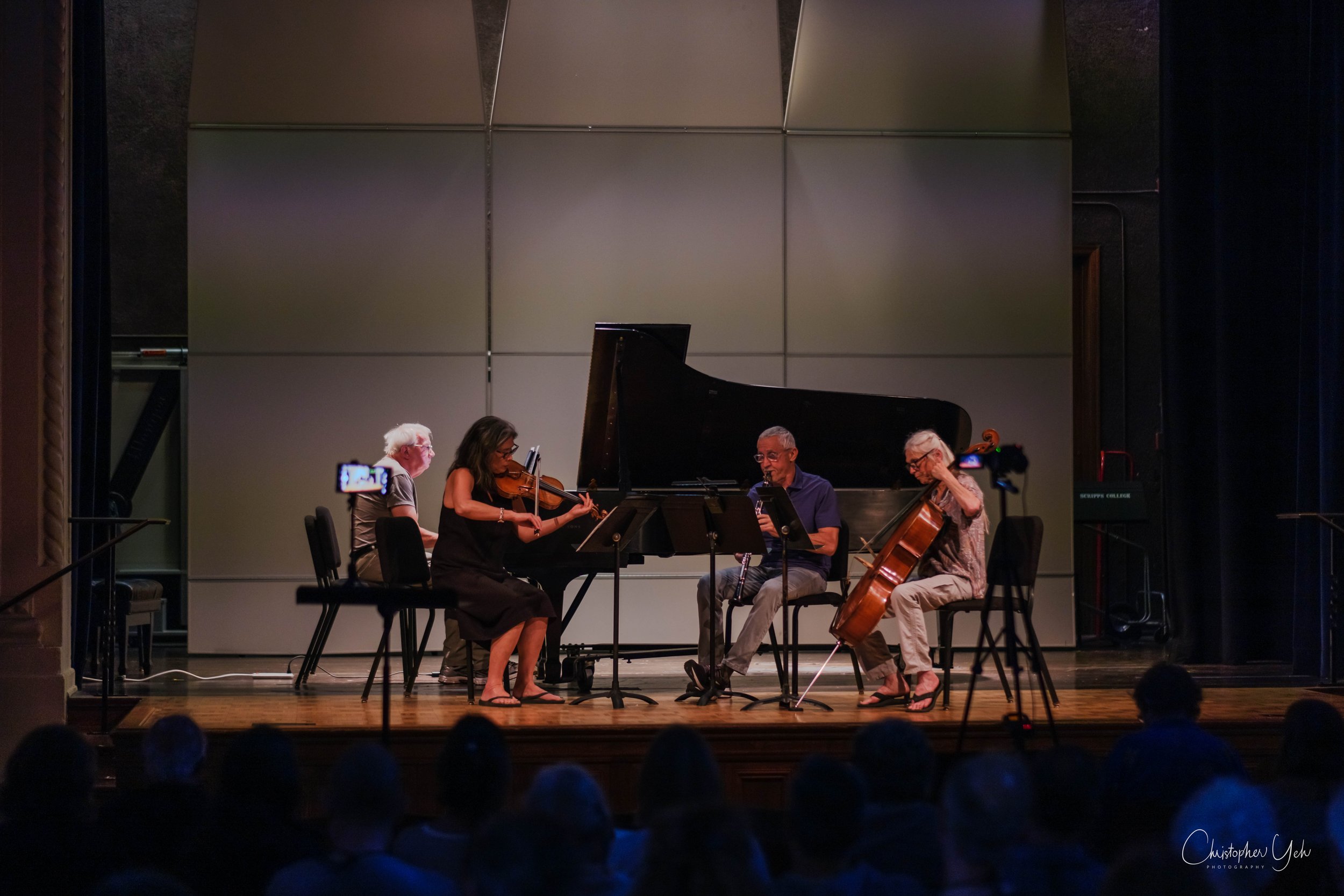 Four musicians performing on stage with a grand piano, two playing violin and one playing cello, in front of an audience at a concert hall.